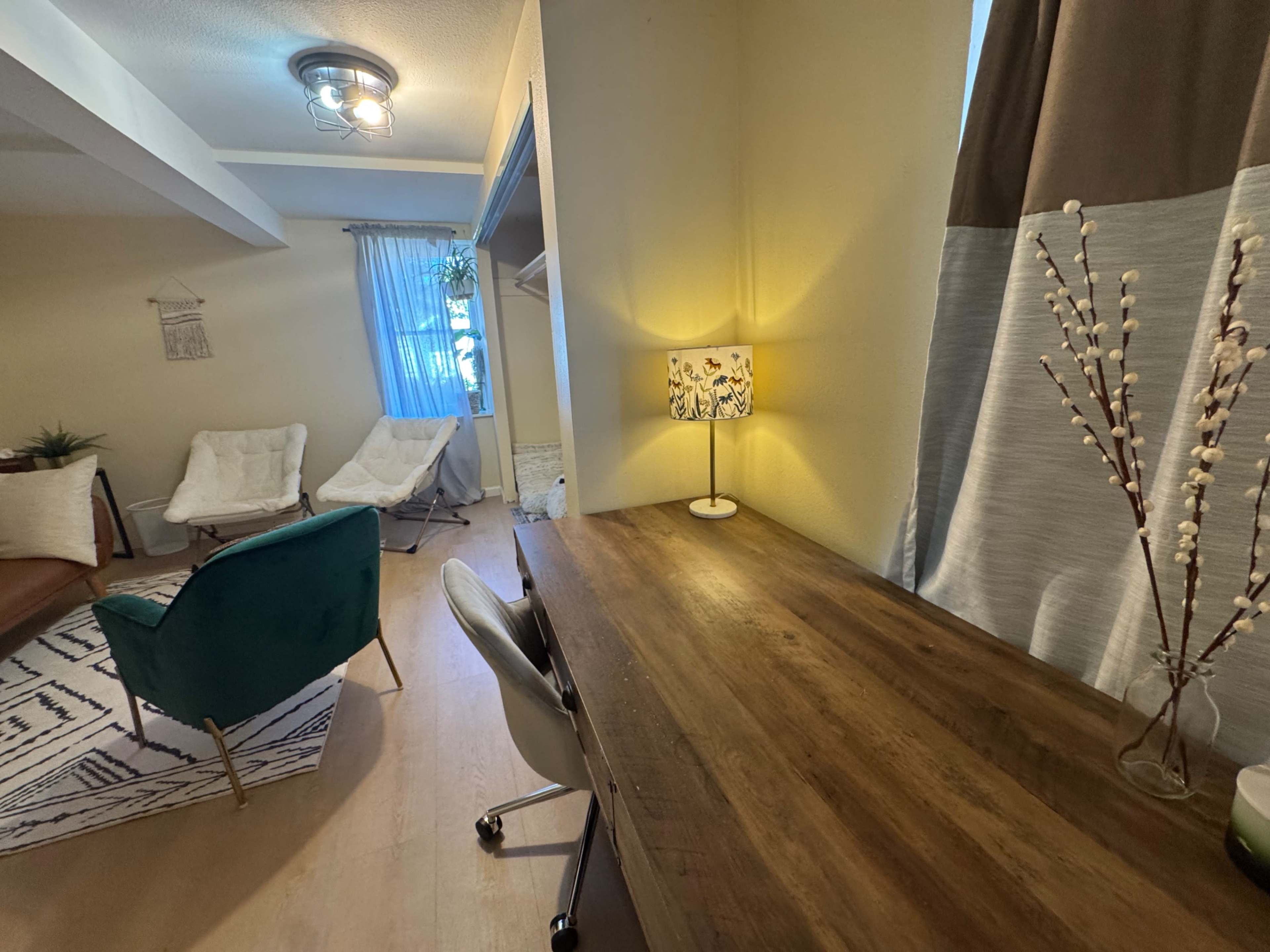 The image shows a cozy interior space featuring a wooden desk, a green chair, two white lounge chairs, a patterned rug, and soft lighting from a lamp.