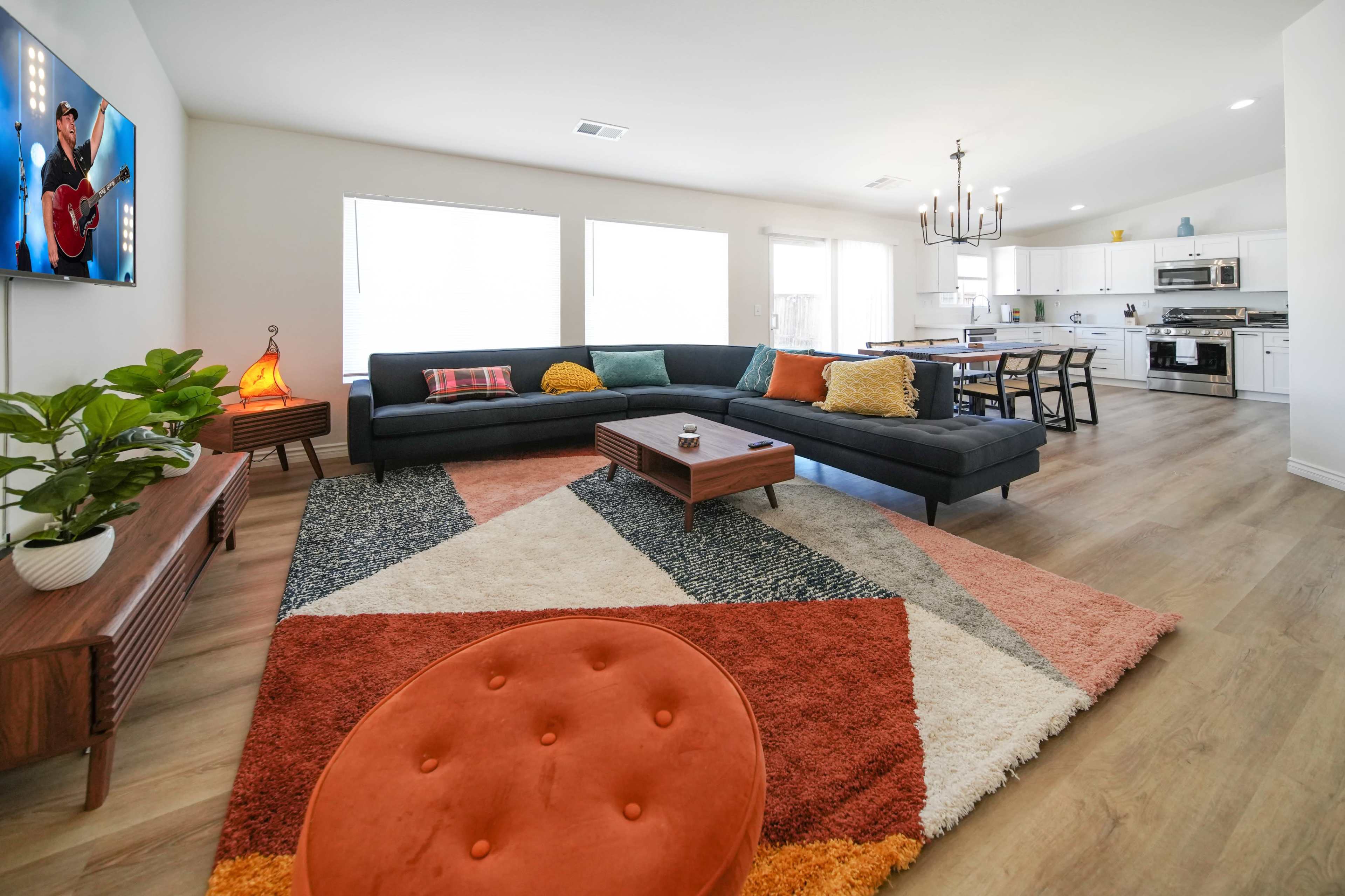 A spacious living room features a large sectional sofa, a colorful geometric area rug, a flat-screen TV, and an adjacent open kitchen.