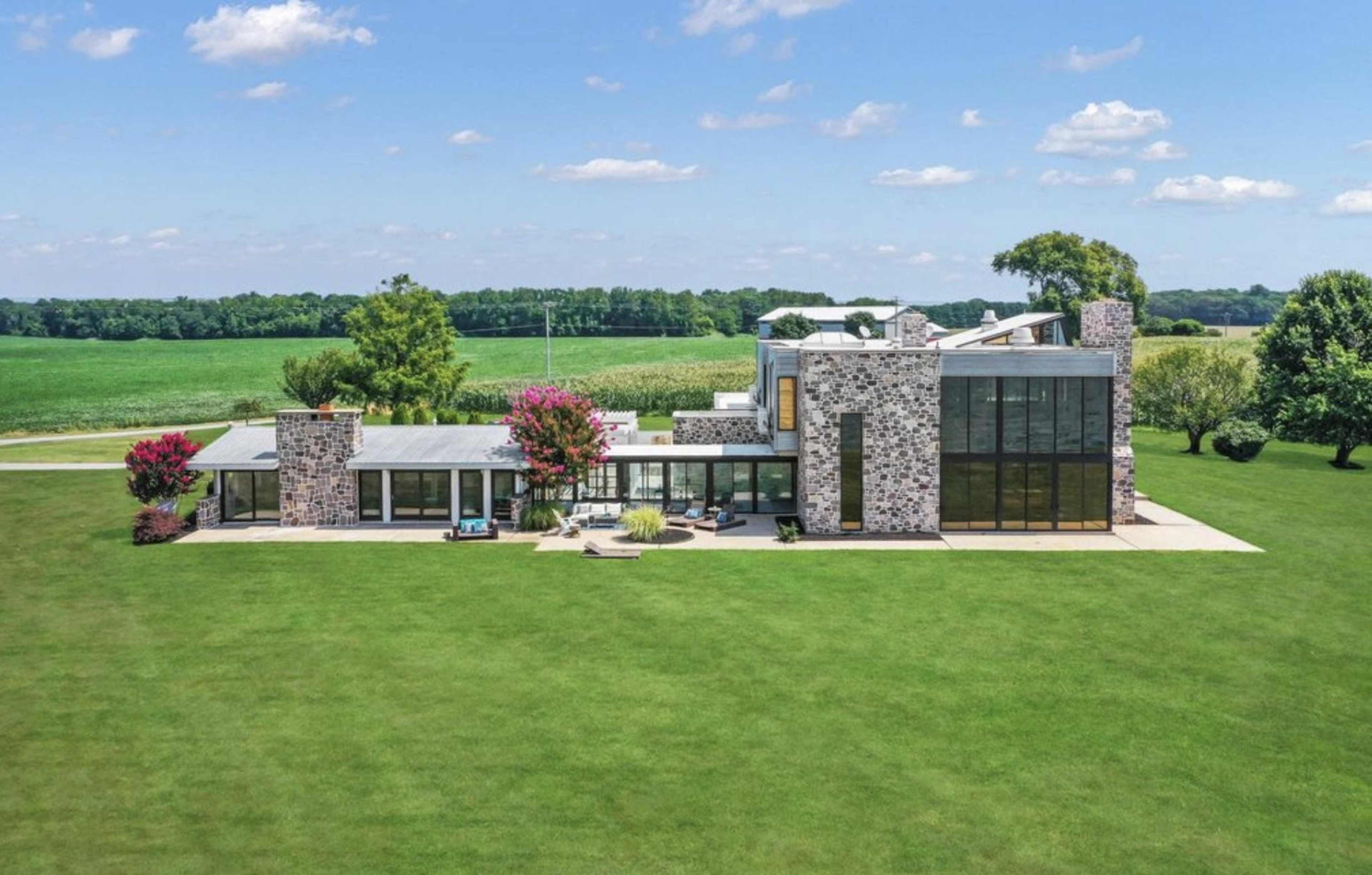 A modern stone and glass house is set on a vast green lawn with trees and open fields in the background.