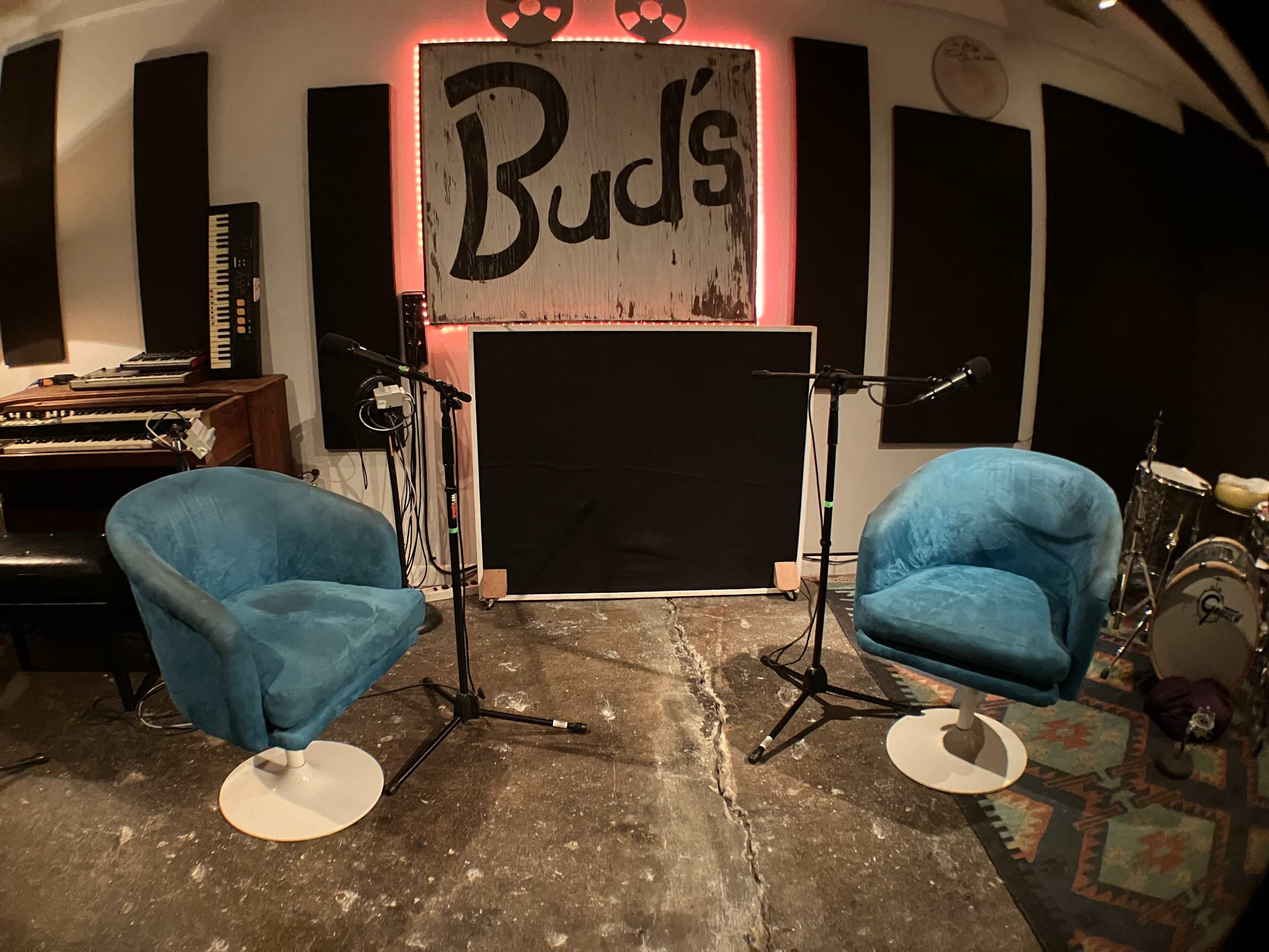 The image shows a studio setting with two blue chairs facing a blank black panel, surrounded by musical instruments and a sign that reads "Bud's."