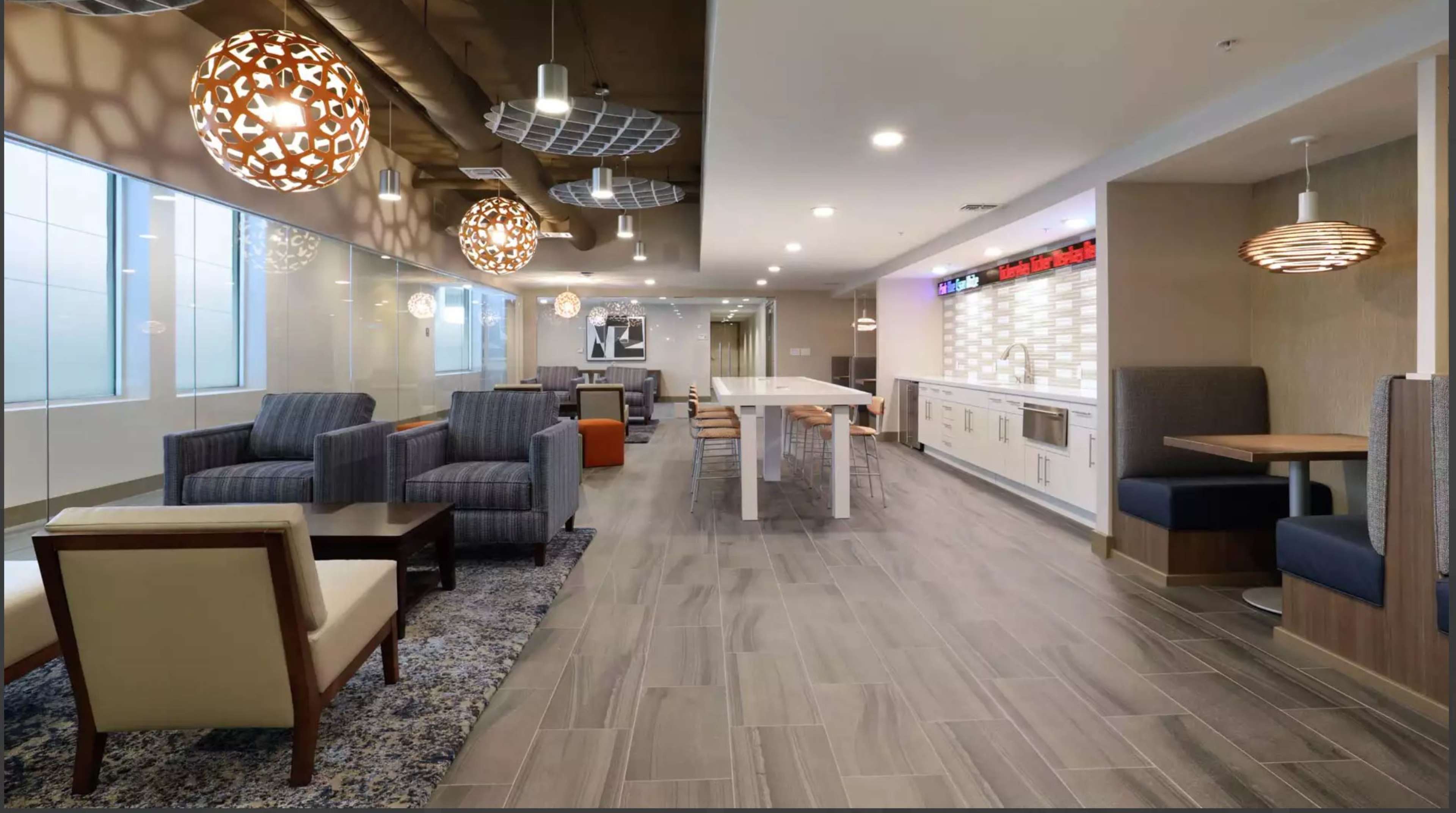 A modern lounge area with seating arrangements, pendant lights, and a kitchenette along one wall.