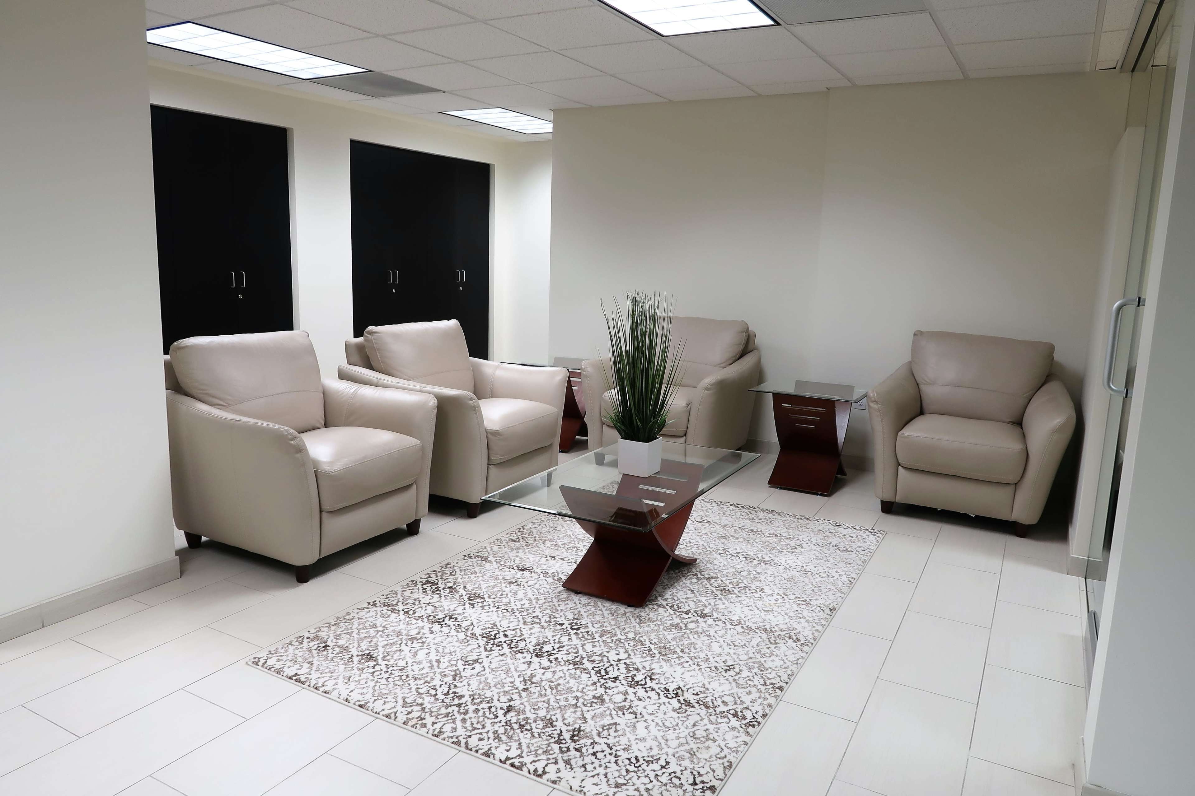 A modern waiting area features beige leather chairs, a glass coffee table, and a decorative rug on a tiled floor.