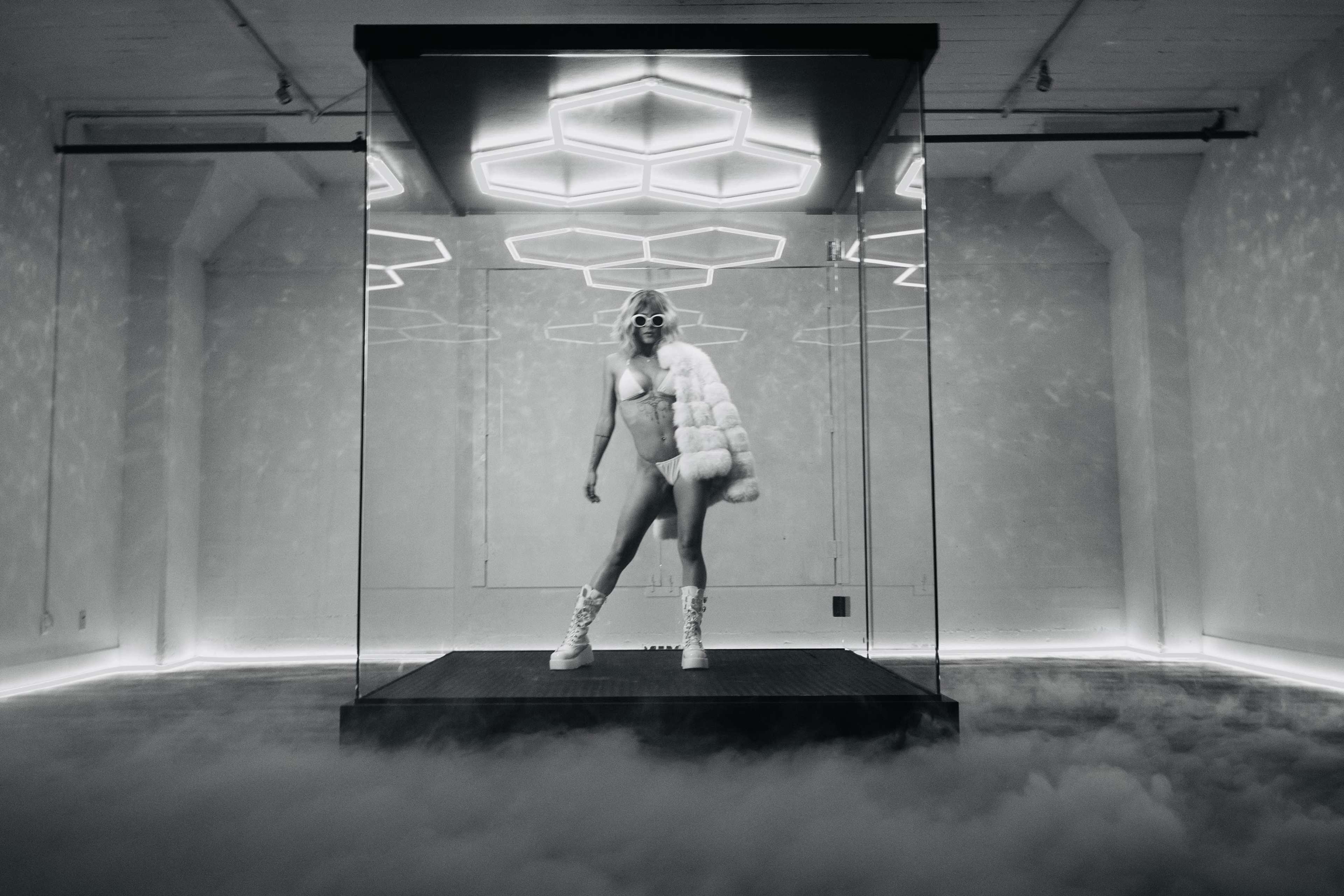 A person in a fur coat and sunglasses poses inside a glass enclosure with geometric light patterns above them.