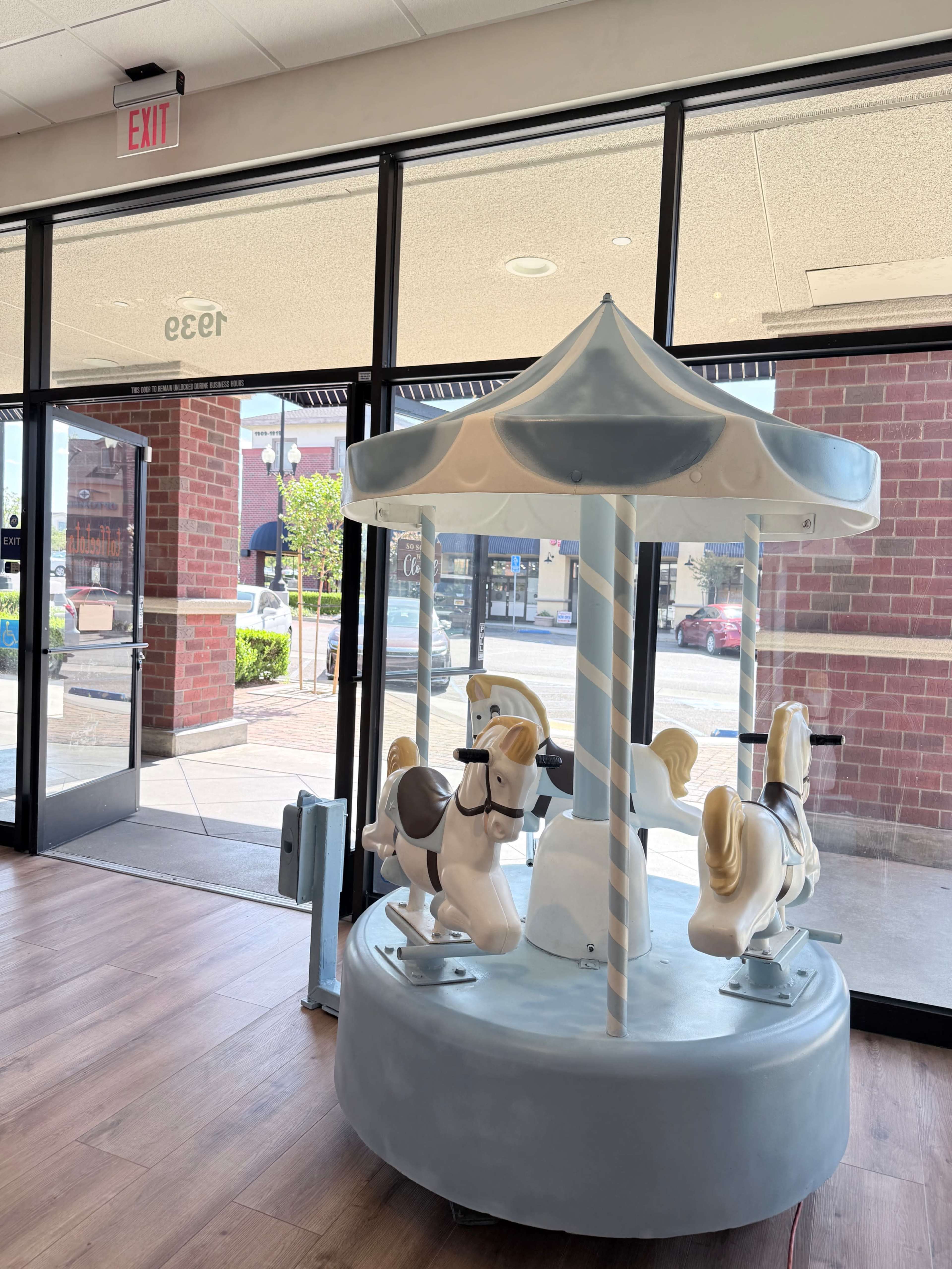 A small carousel with horse figures is placed inside a store, visible through large glass doors leading to the outside.