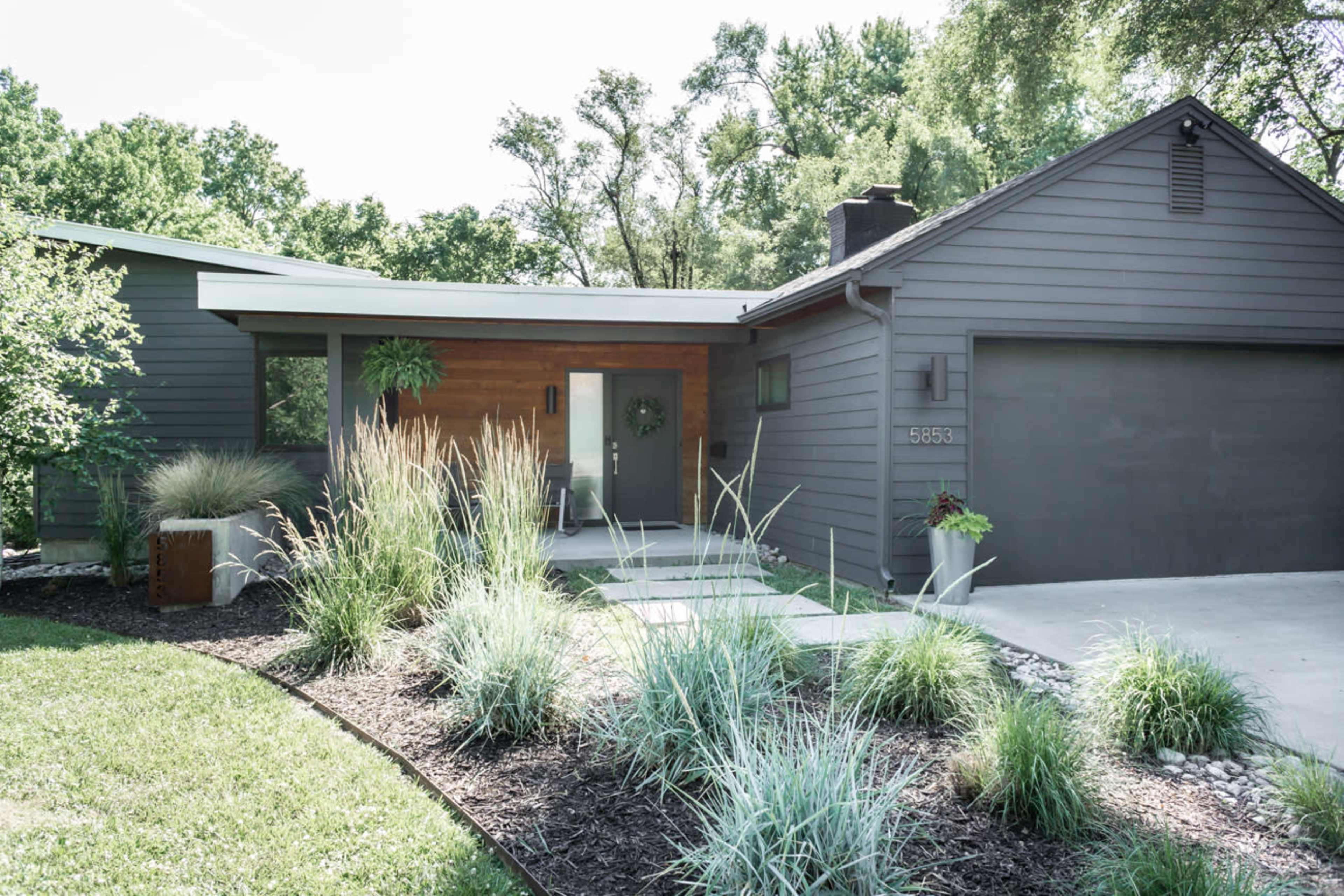 A modern house with a combination of dark gray siding and natural wood accents, featuring a landscaped front yard with ornamental grasses and a concrete walkway.