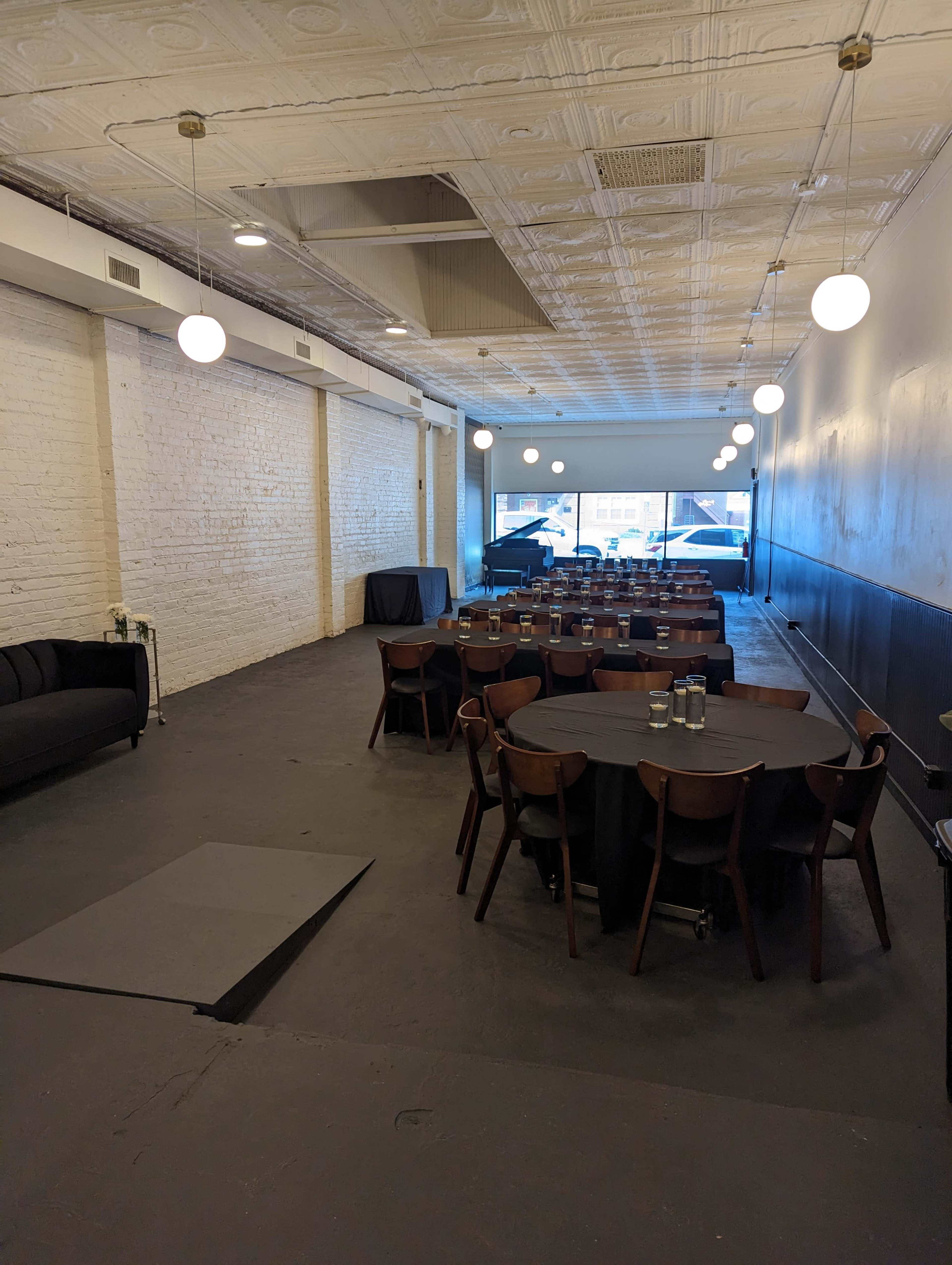 A spacious, modern event venue with round tables set for a gathering, a black couch in the corner, and large windows at the far end.