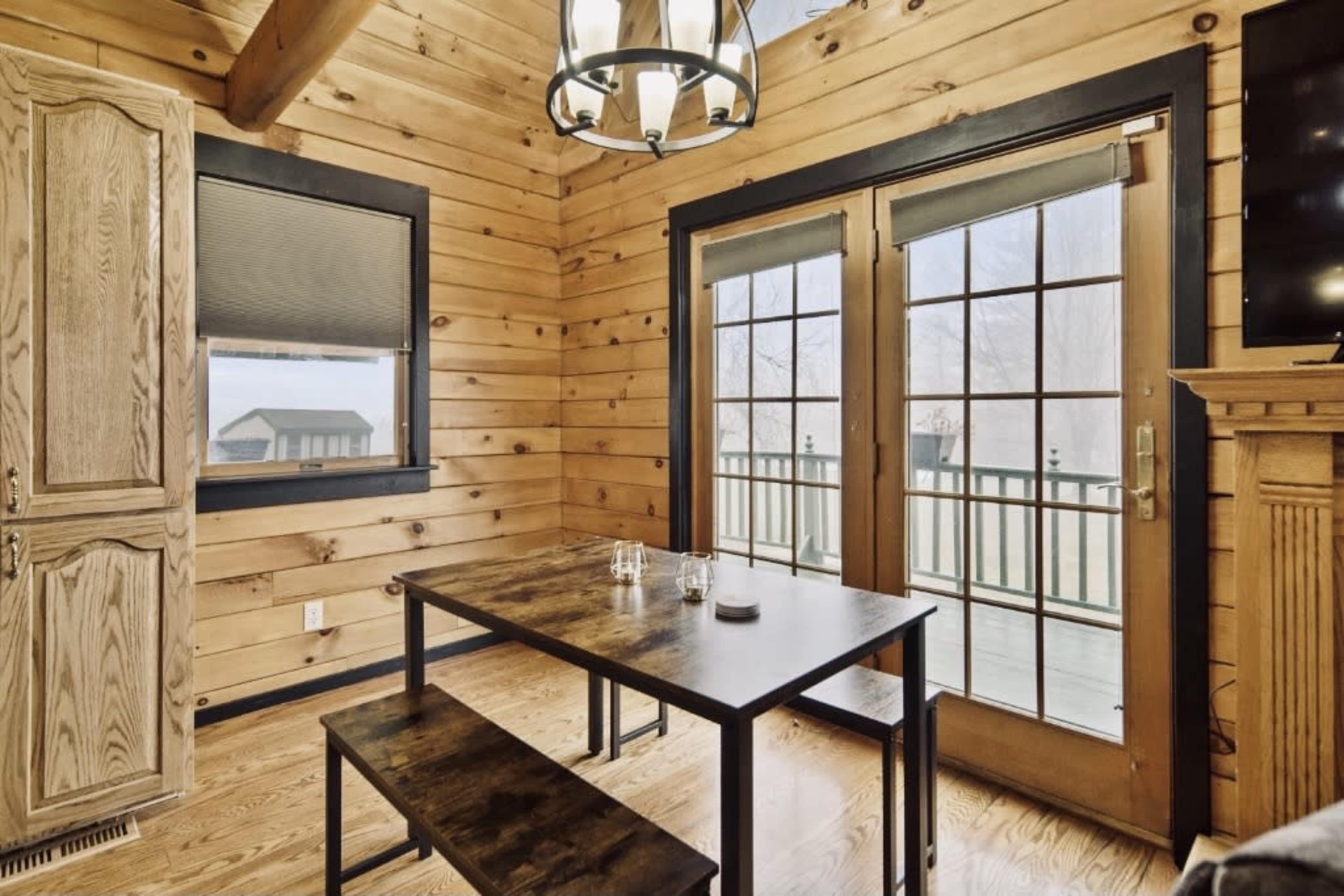 A dining area features a wooden table and benches, with large double doors opening to a deck and wooden paneled walls.
