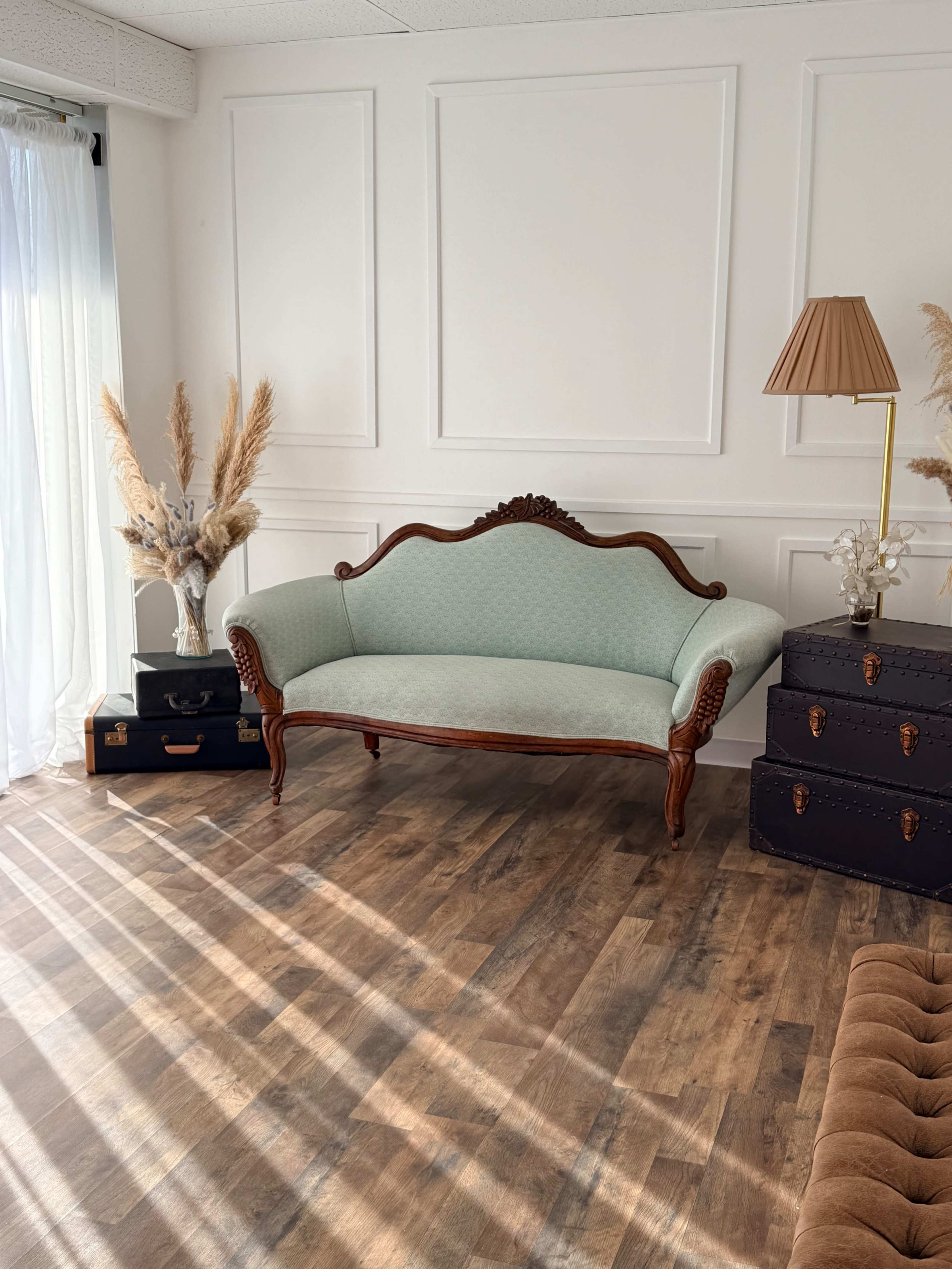 The image features a vintage-style blue upholstered loveseat positioned near a bright window, accompanied by a decorative lamp and potted plants, all set against a wall with paneling.