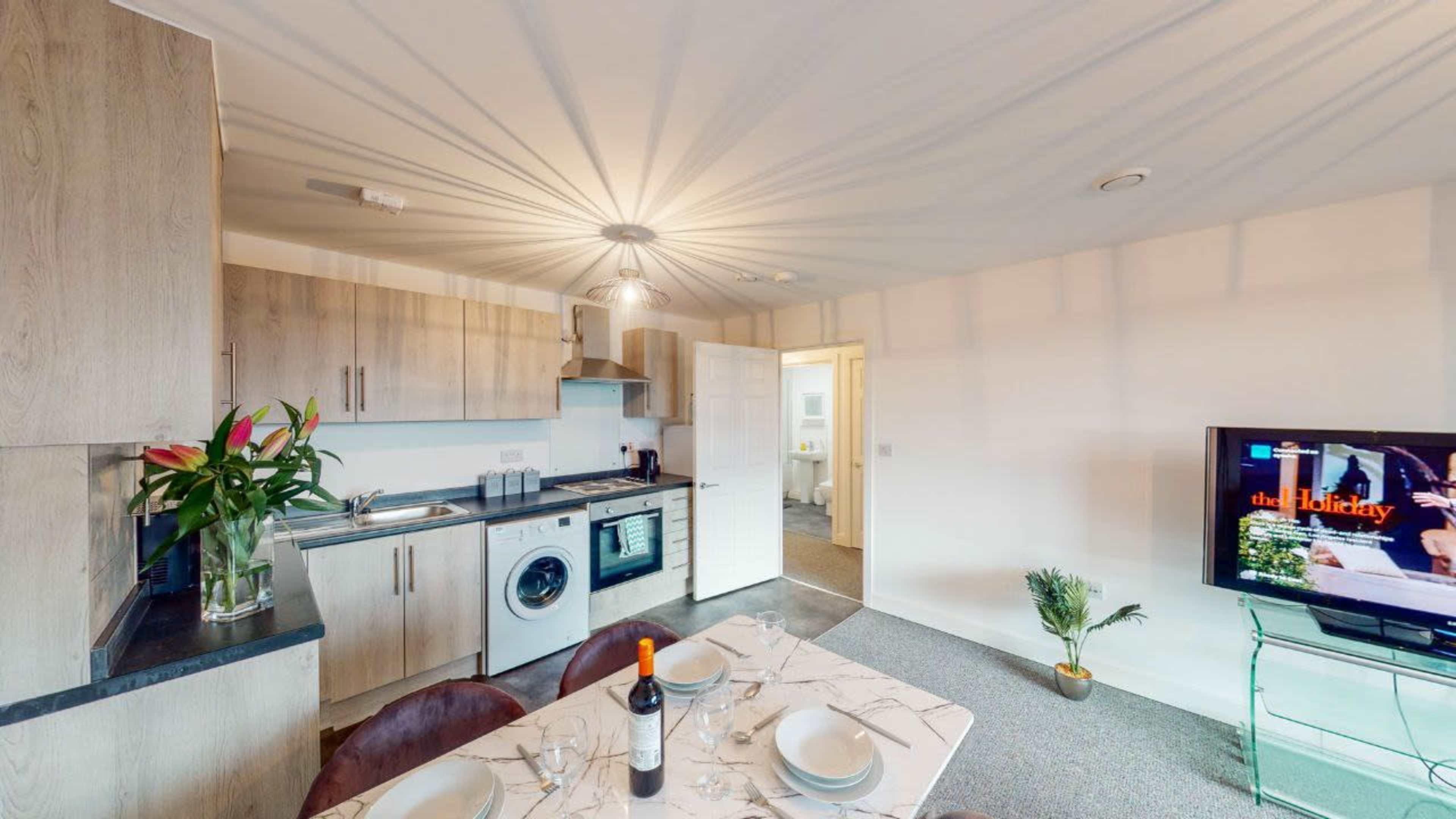 A modern kitchen and living area feature wooden cabinets, a dining table set for dinner, and a television displaying a movie.