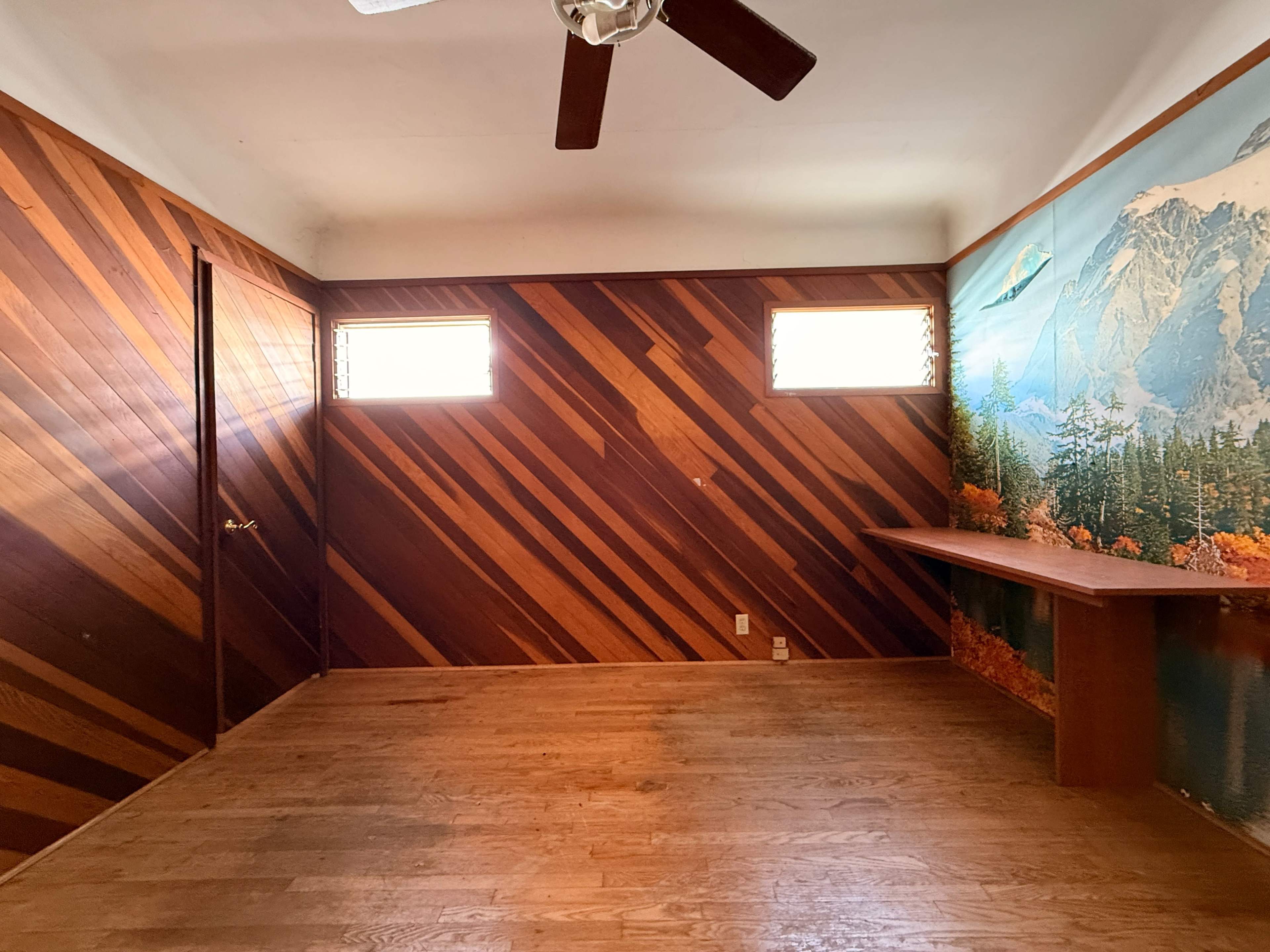 The image shows an empty room with wooden diagonal wall panels and a mural of a mountain landscape on one wall.