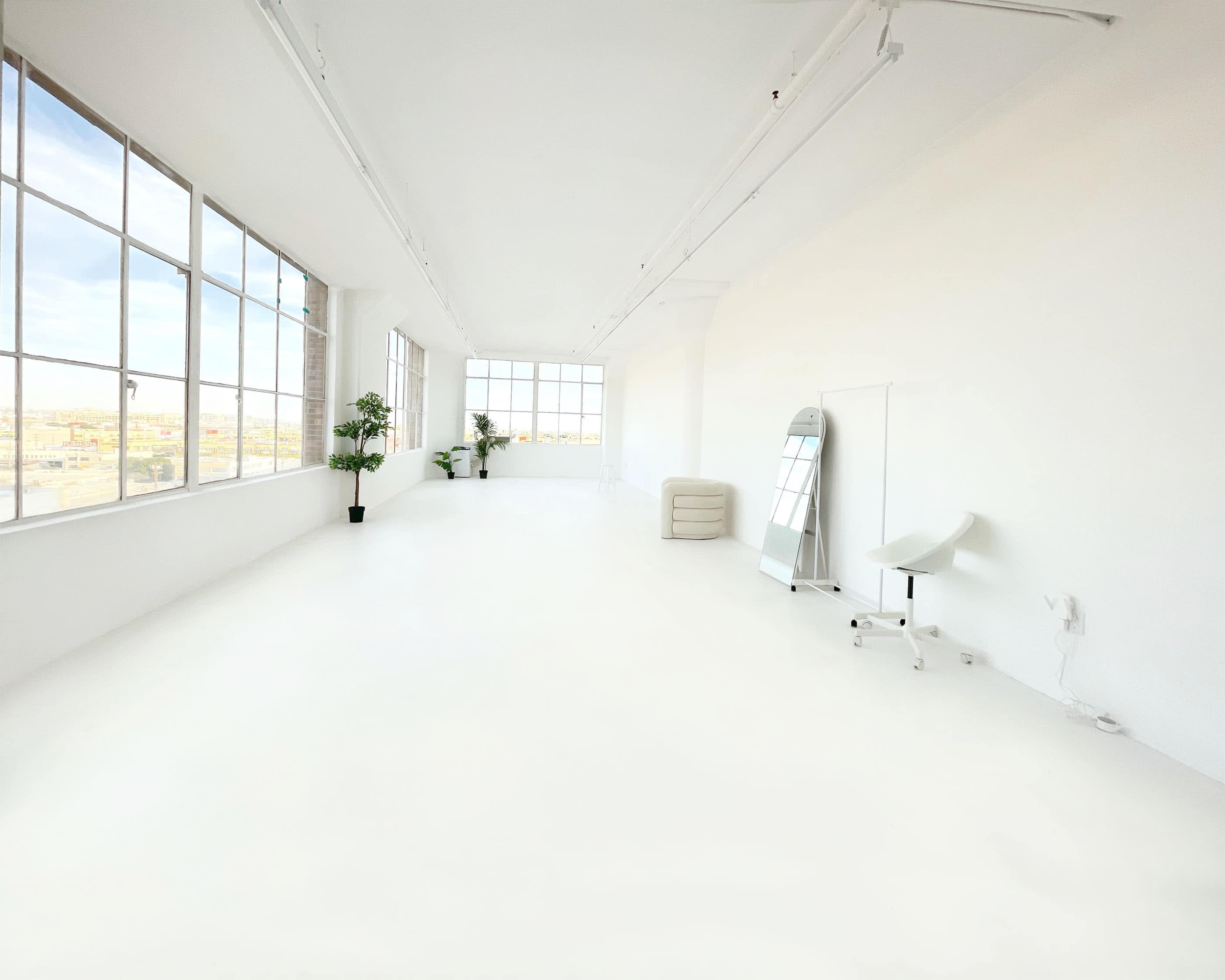 A spacious, minimalistic room with large windows, white walls, and a few pieces of furniture, including a mirror and a chair.
