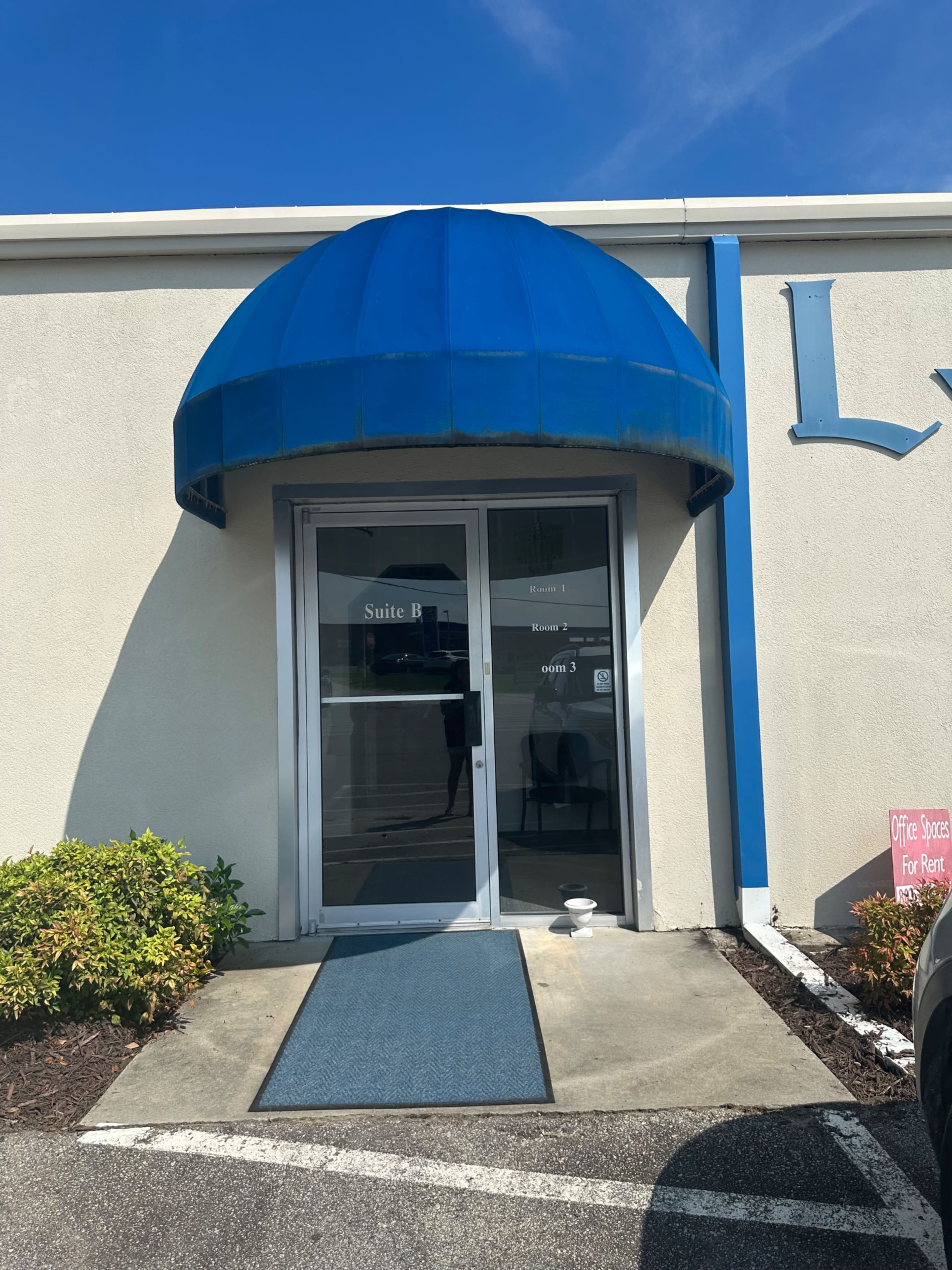 The entrance of a commercial building features a blue awning above double glass doors, with a blue mat leading to the entrance.