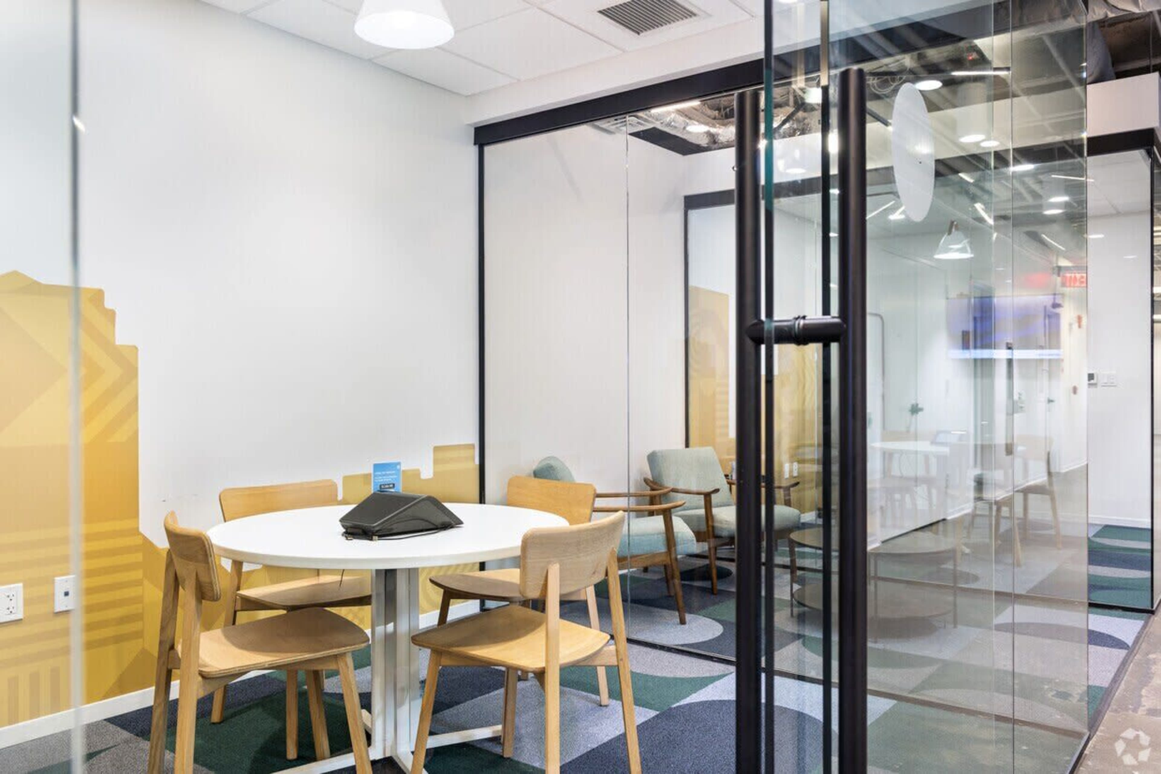 The image shows a glass-walled conference room with a round table, four wooden chairs, and a TV mounted on the wall.