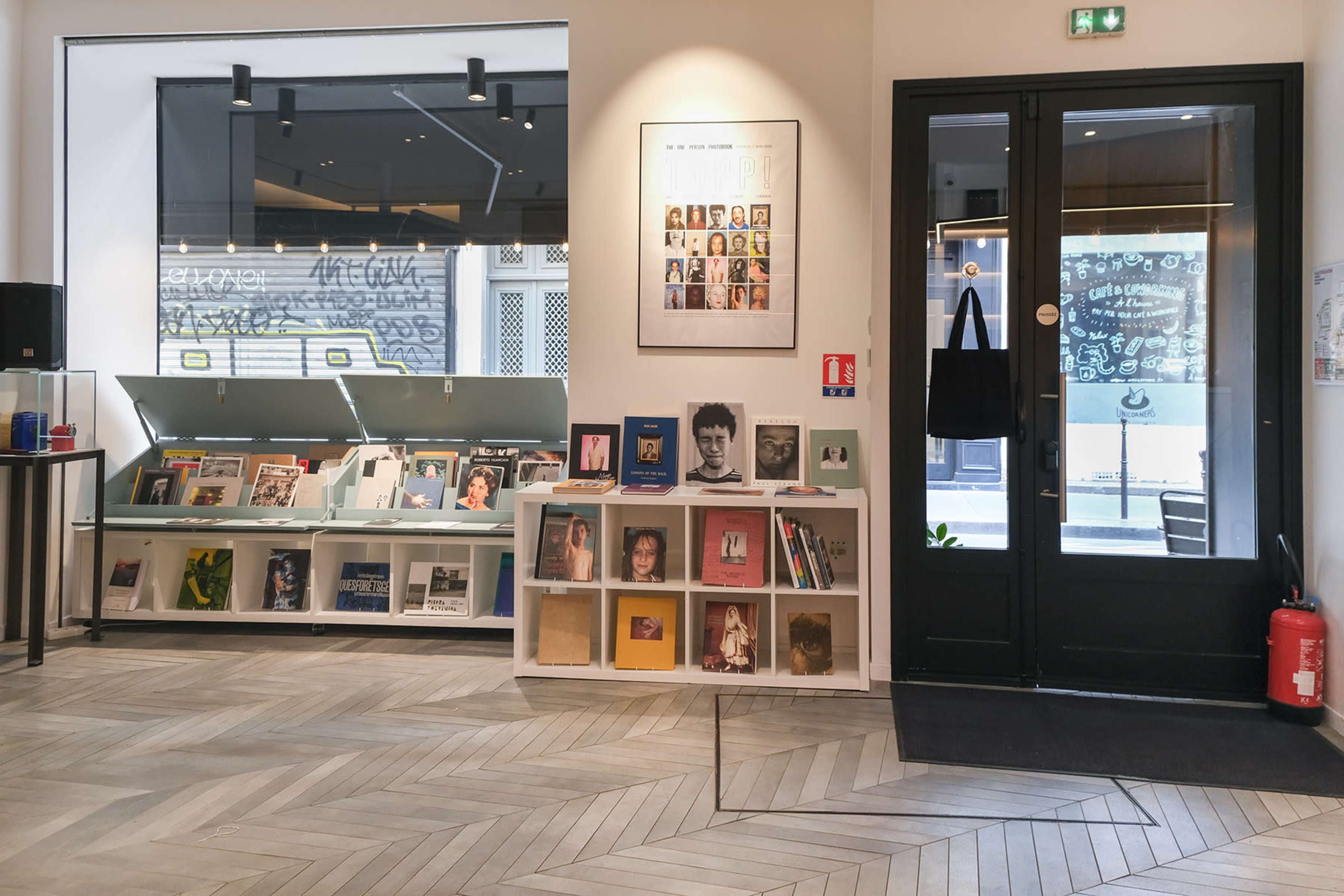 The image shows a modern interior of a bookstore or gallery featuring shelves of books and photographs on the left, and a glass entrance door on the right.