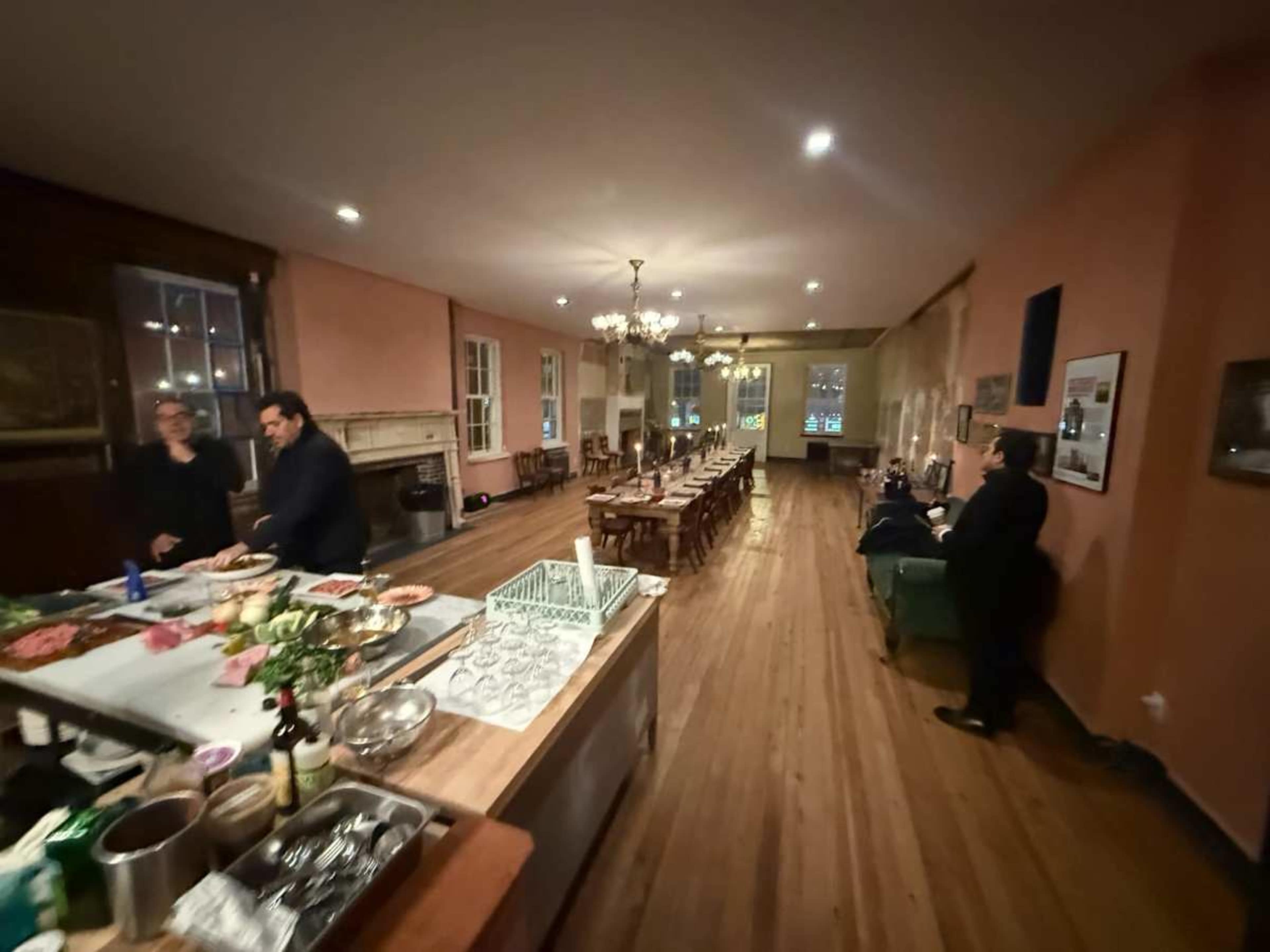 A spacious dining room features a long table set for a meal, with two people preparing food in the kitchen area on the left.