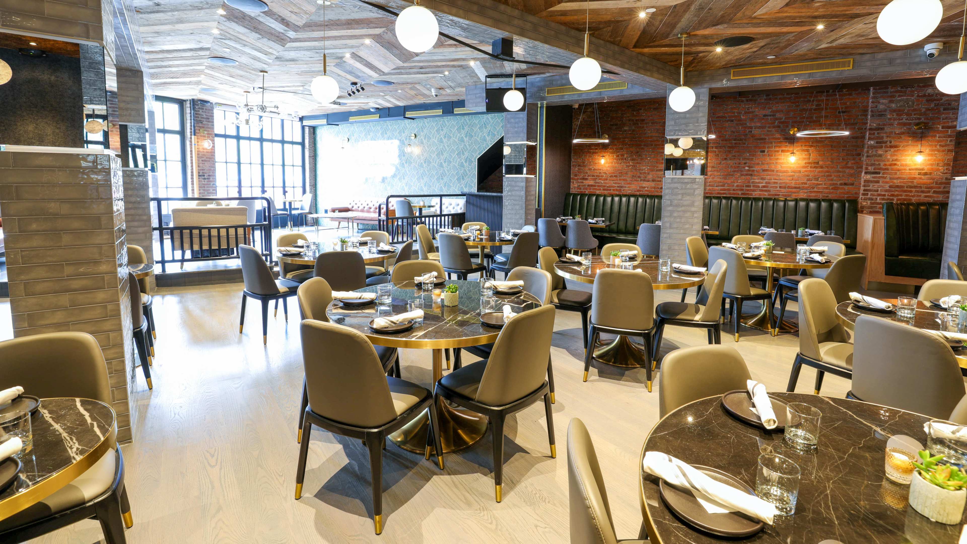 The image shows a modern restaurant interior featuring round tables with dark marble tops, gray chairs, and large pendant lights, set against a backdrop of exposed brick and wooden ceiling details.
