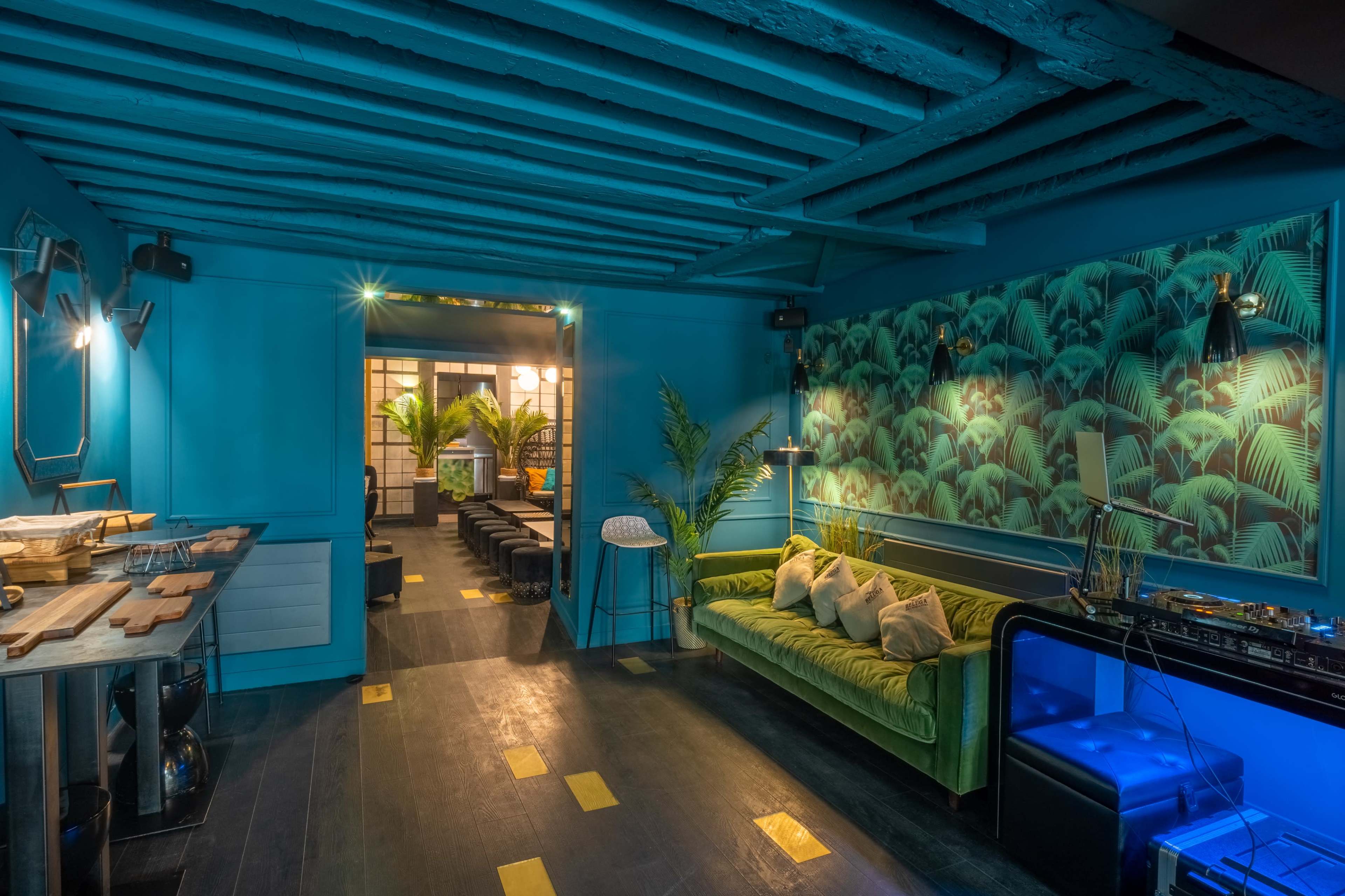 A vibrant blue room with a green velvet sofa, palm leaf wallpaper, and wooden beams on the ceiling, leading to an illuminated entryway.