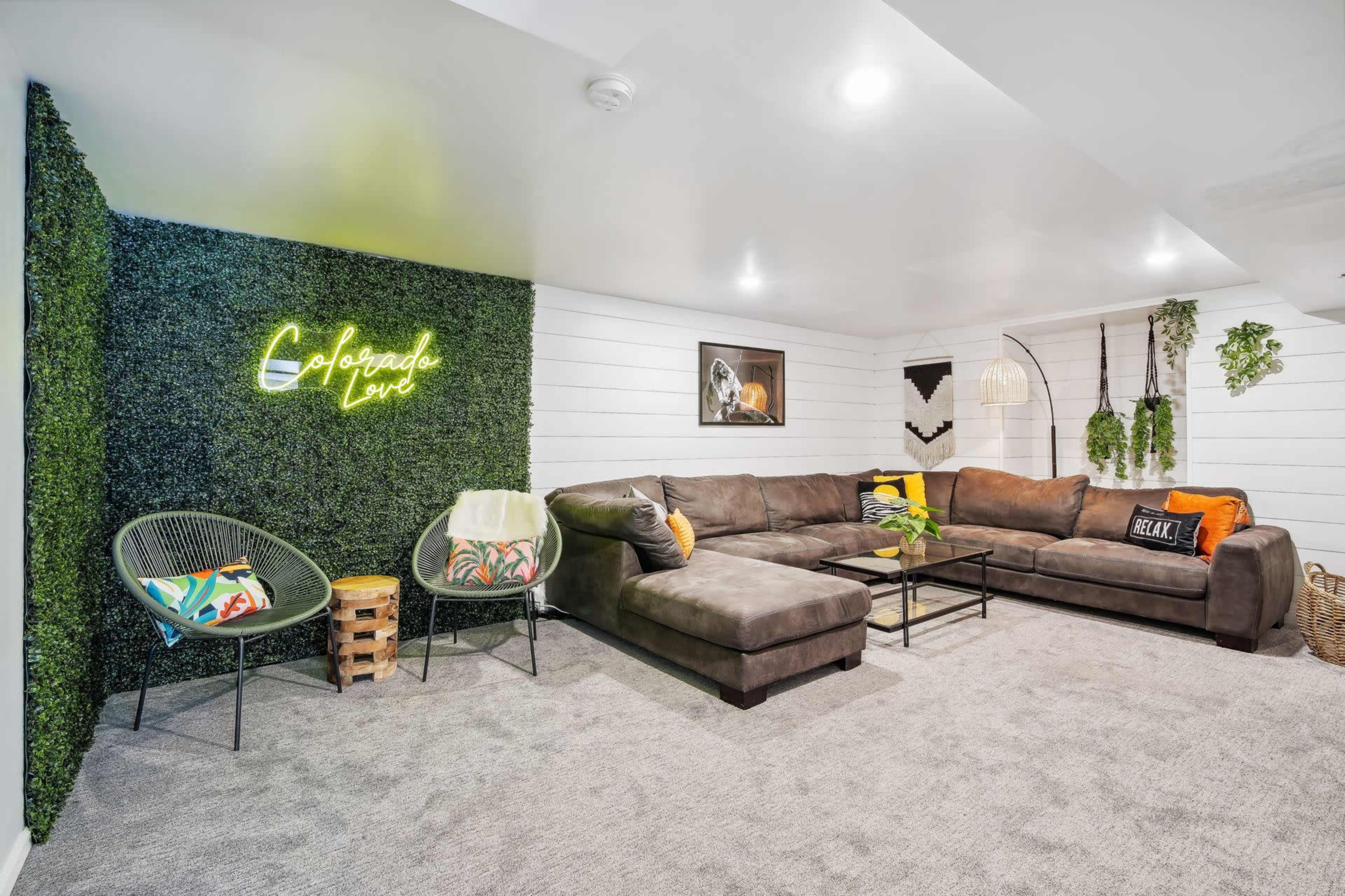 A modern basement living room with a large sectional sofa, a neon sign reading "Colorado Love," and a green wall with faux greenery.