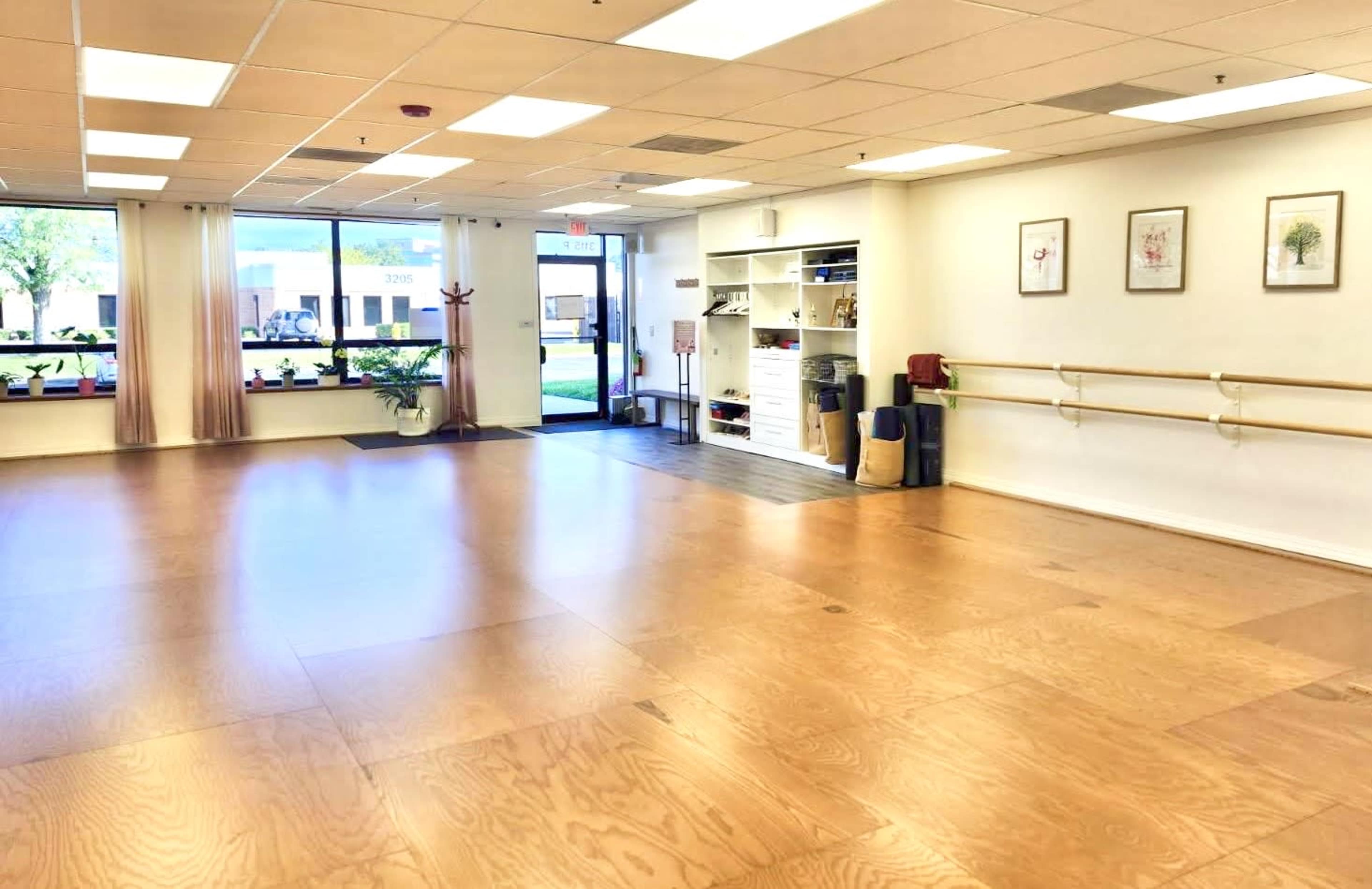 The image shows an empty dance studio with wooden flooring, mirrored walls, and large windows allowing natural light to enter.
