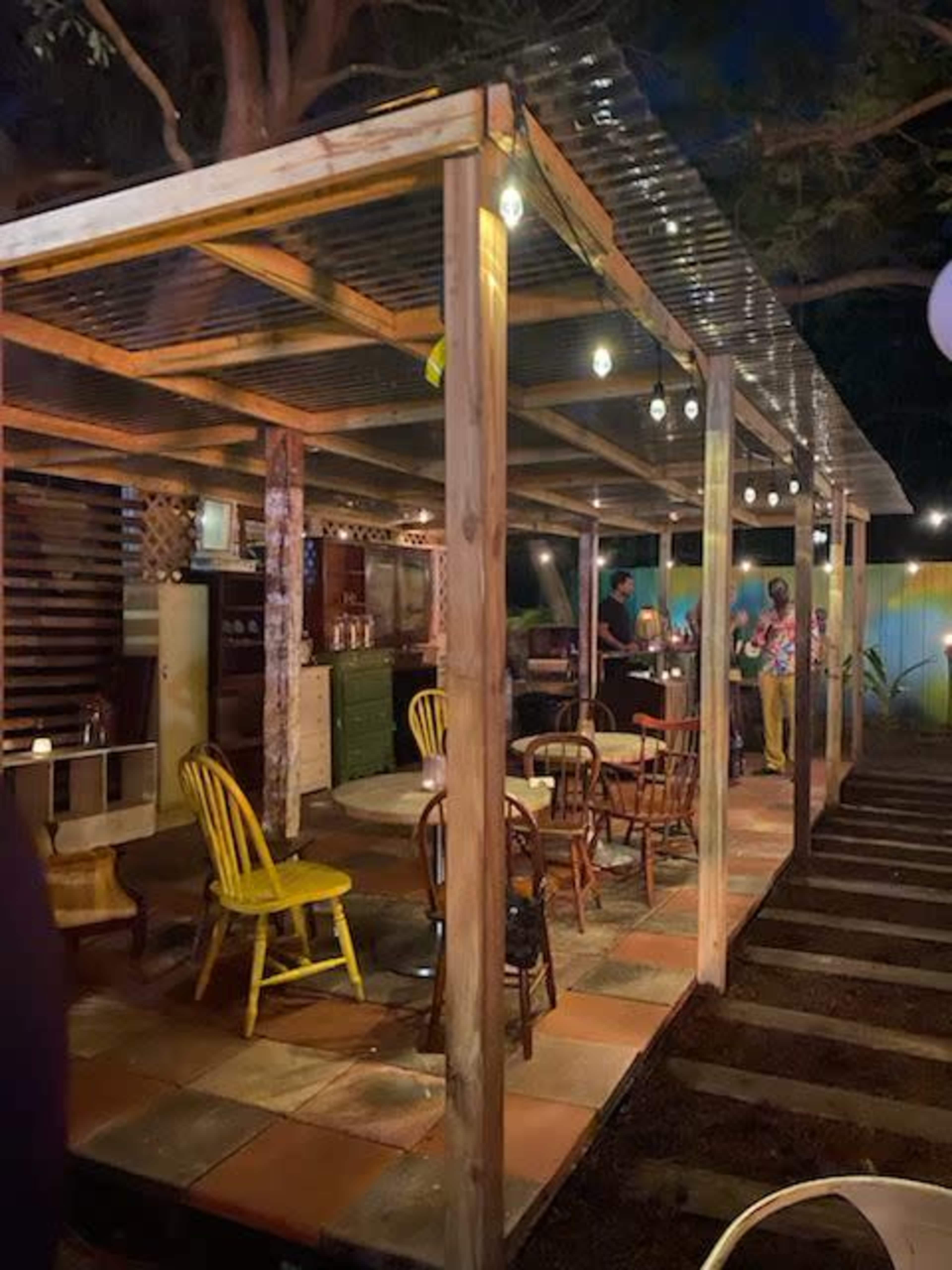 A rustic outdoor dining area with wooden chairs and tables under a transparent roof, illuminated by string lights in a nighttime setting.