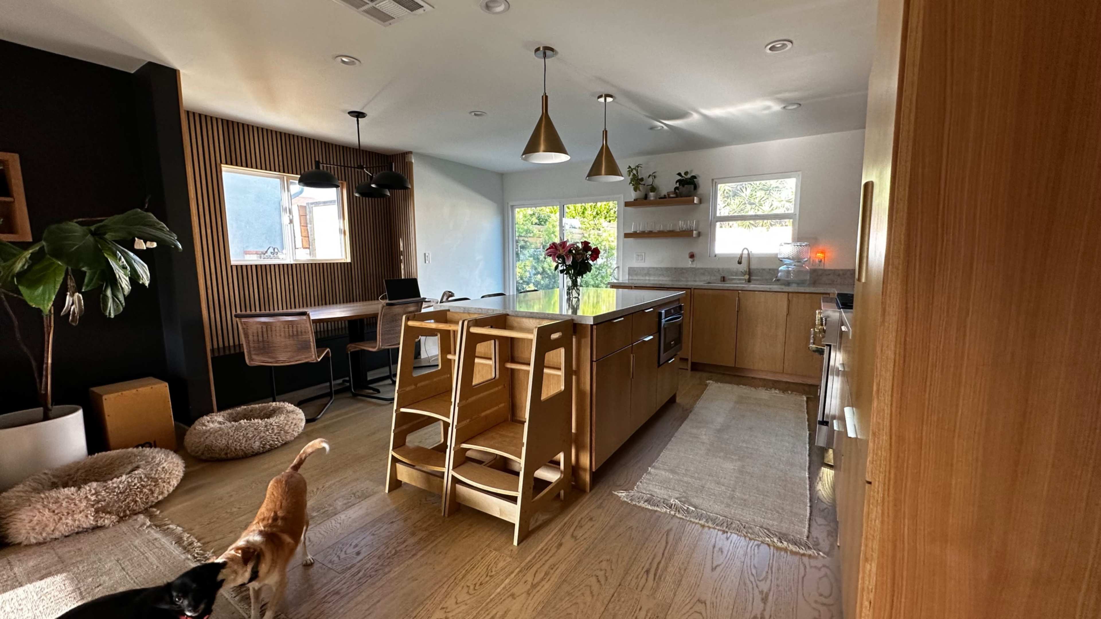A modern kitchen features wooden cabinetry, a central island with seating, and a workspace adjacent to a large window, alongside plush pet beds and two dogs in the scene.