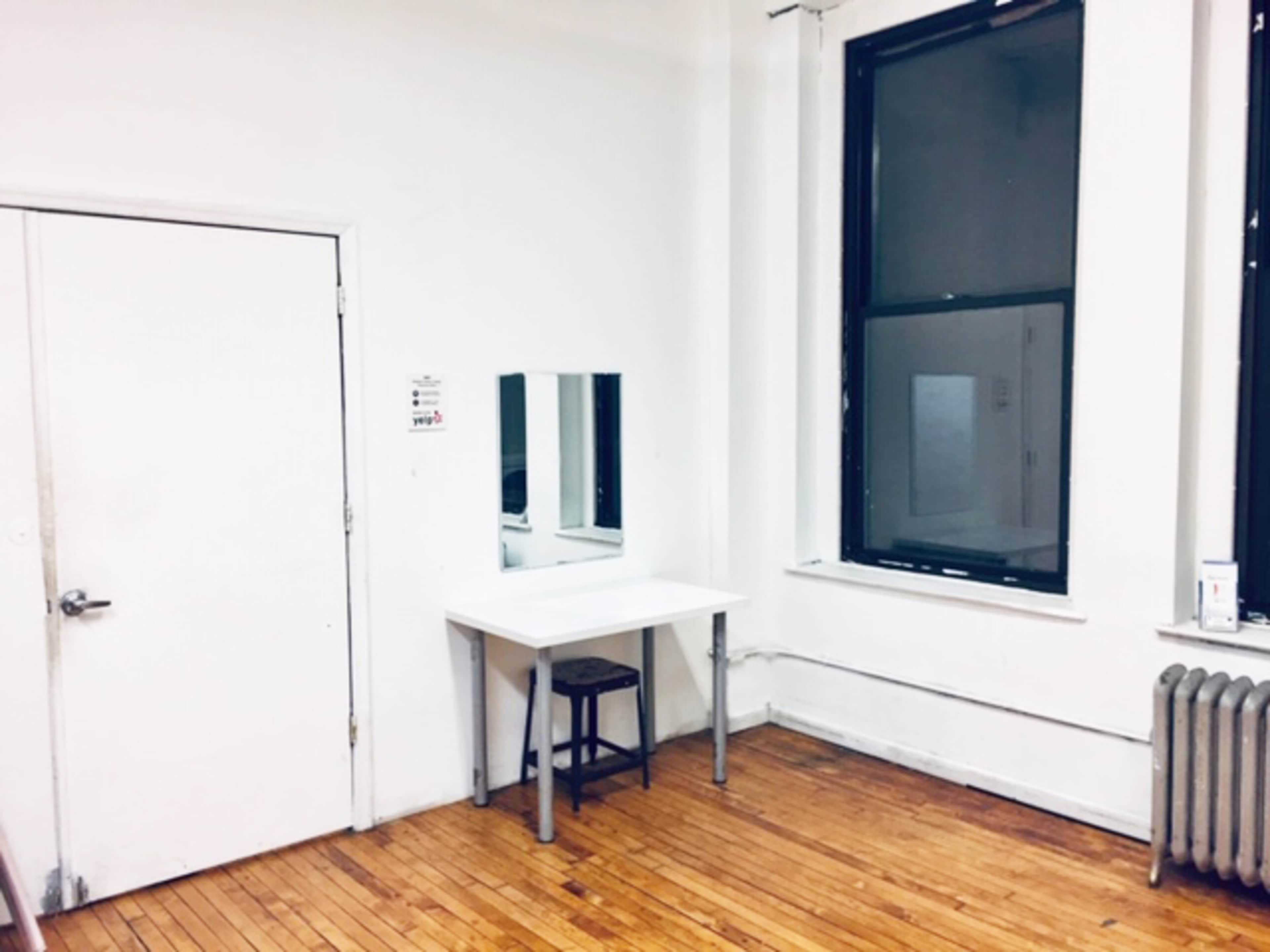 A small, sparsely furnished room with a white wall, a wooden floor, a mirror mounted on the wall above a table, a stool, a window, and a radiator.
