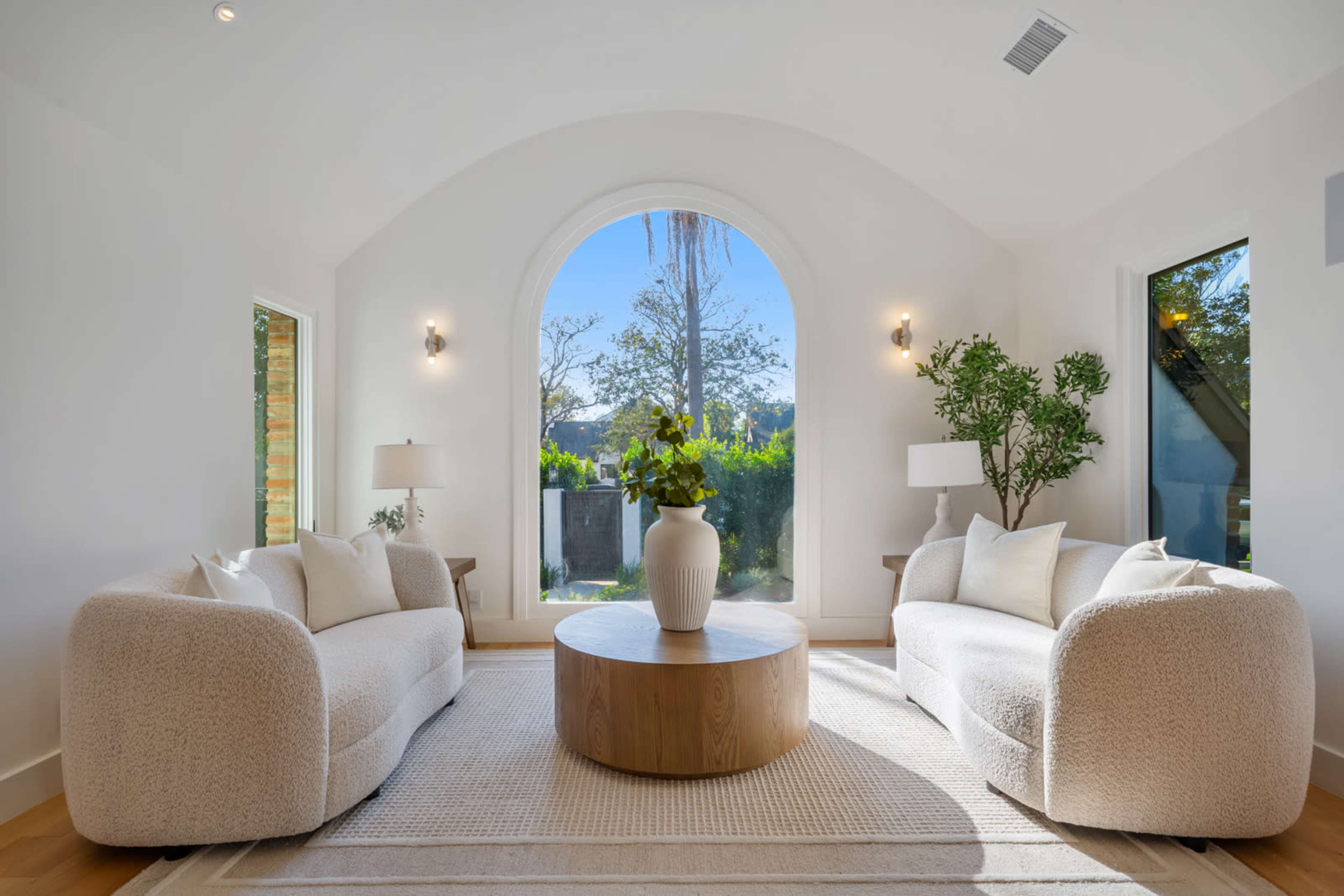A bright, airy living room features two upholstered sofas facing a round wooden coffee table, with large windows providing a view of the outdoors.
