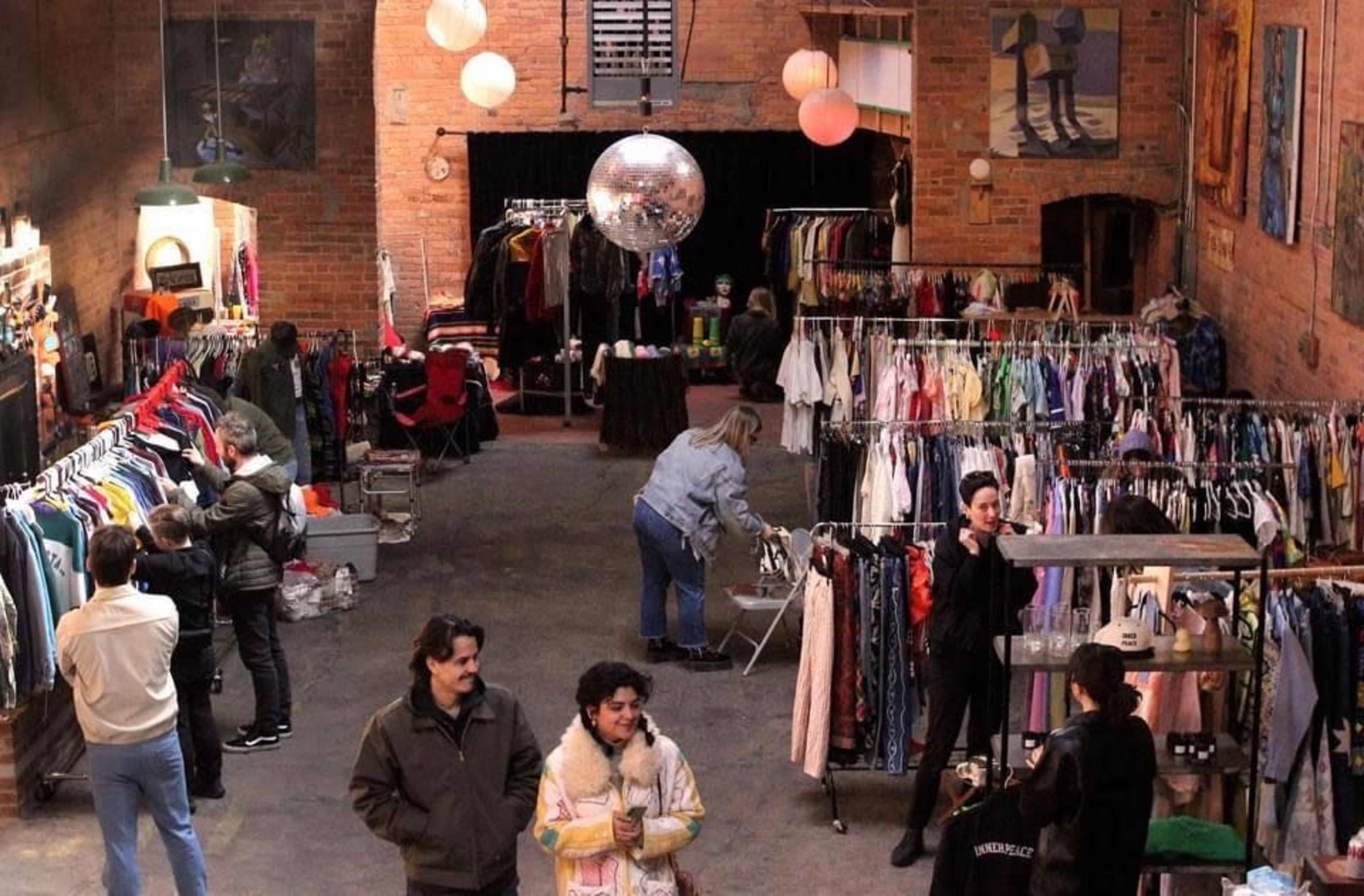 A vintage clothing market features racks of assorted garments, with shoppers browsing and socializing in a spacious warehouse setting.