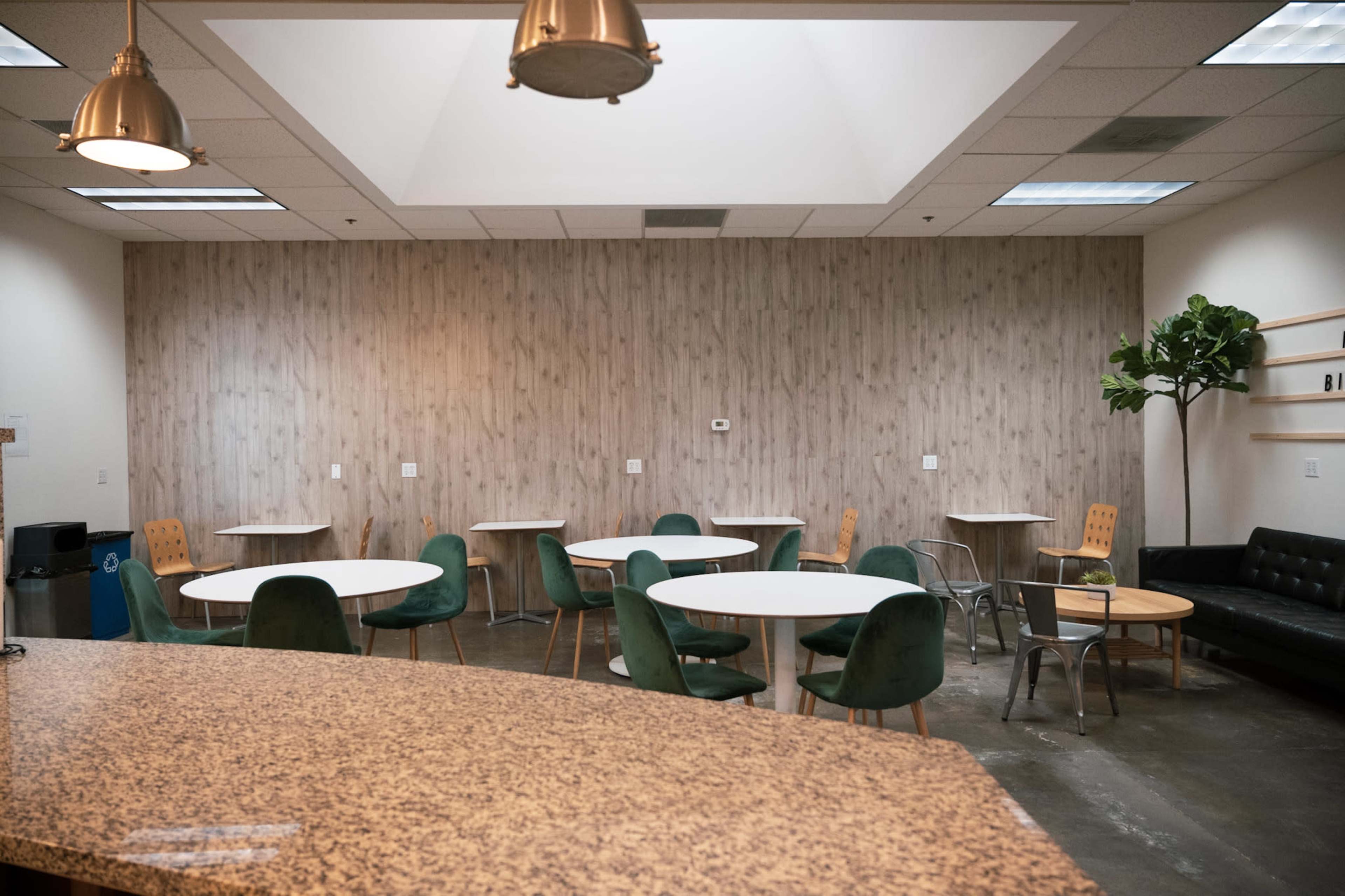 A modern break room with several round tables and chairs, a granite counter, and a large skylight overhead.