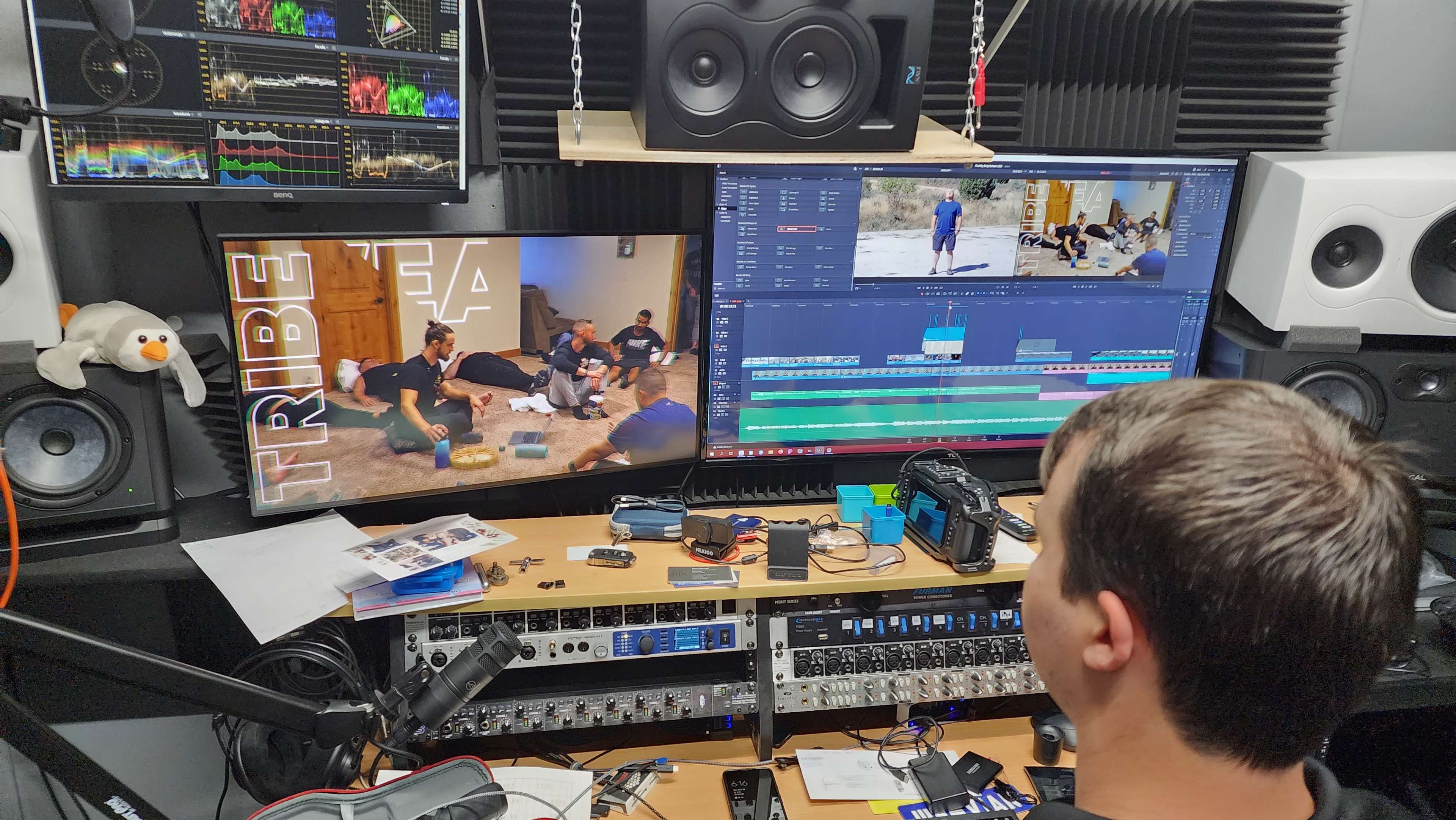 A person is sitting at a desk in a sound studio, working on video editing with dual monitors displaying editing software and footage of a group of people.