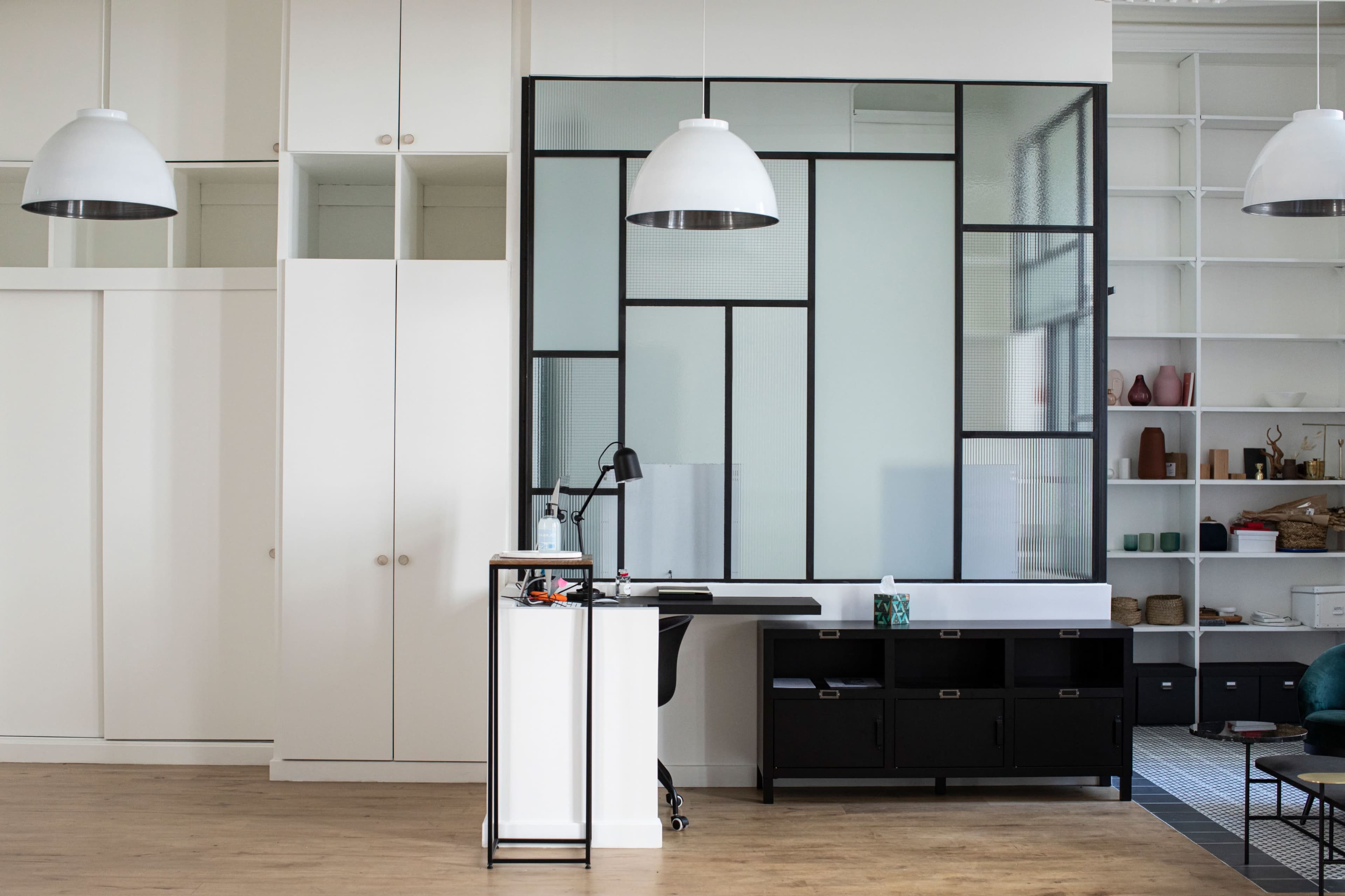 A modern office interior featuring a black desk and a partition with geometric glass panels, surrounded by white cabinetry and shelving.