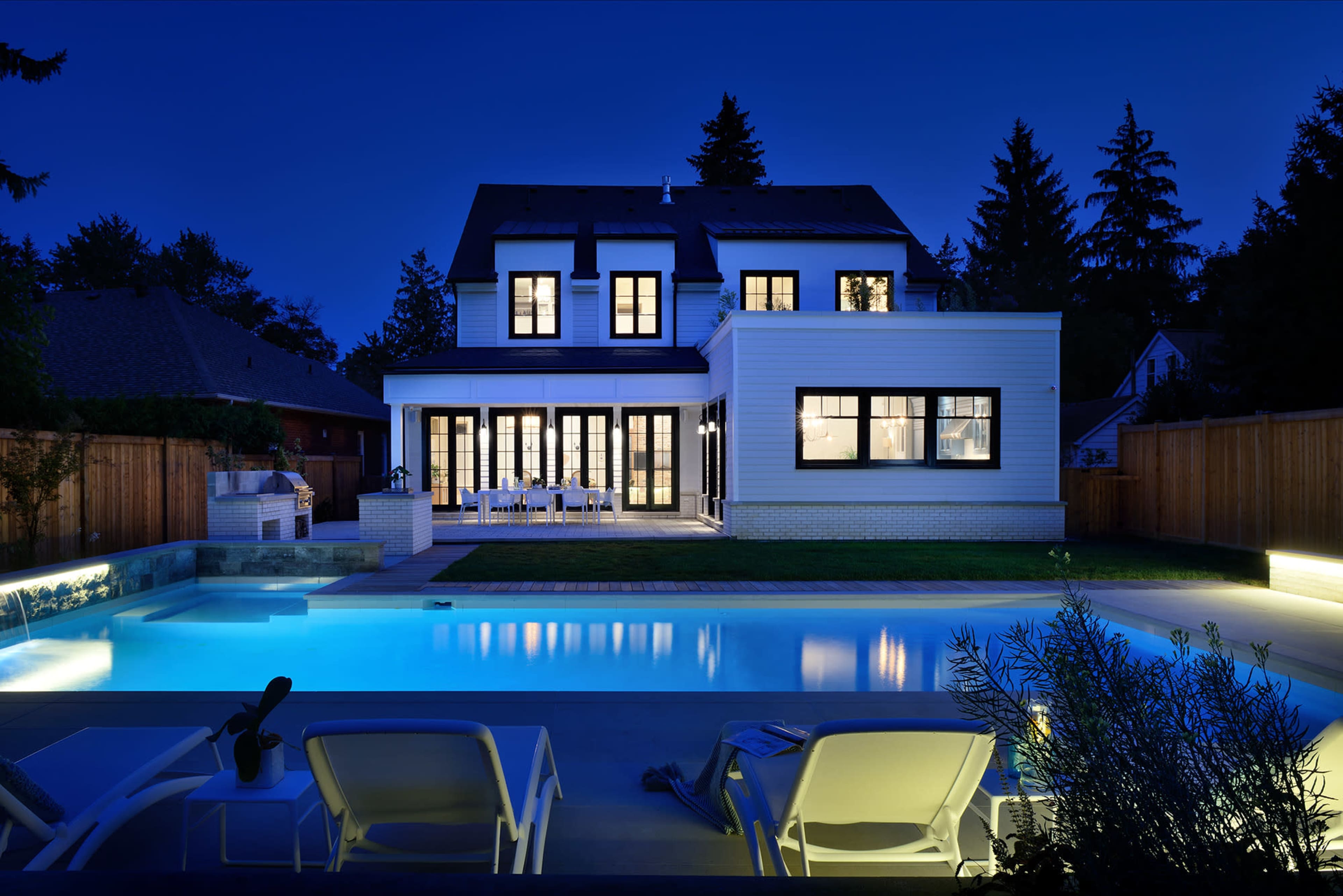 A modern house with large windows and a spacious patio overlooks a well-lit pool at night.