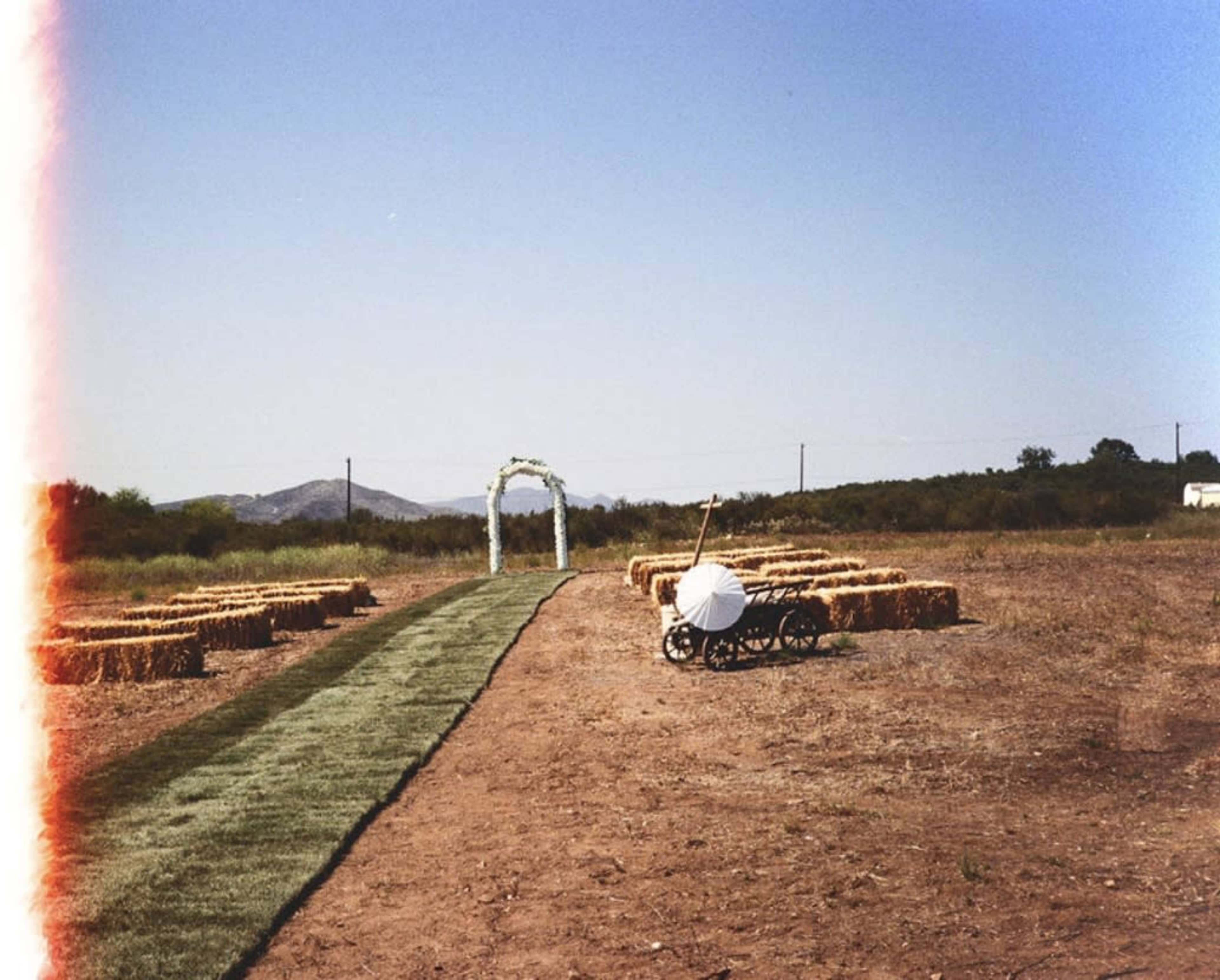 An outdoor setting features a grassy aisle leading to an archway, flanked by bales of hay arranged in a semi-circle, with a wheelbarrow positioned nearby.