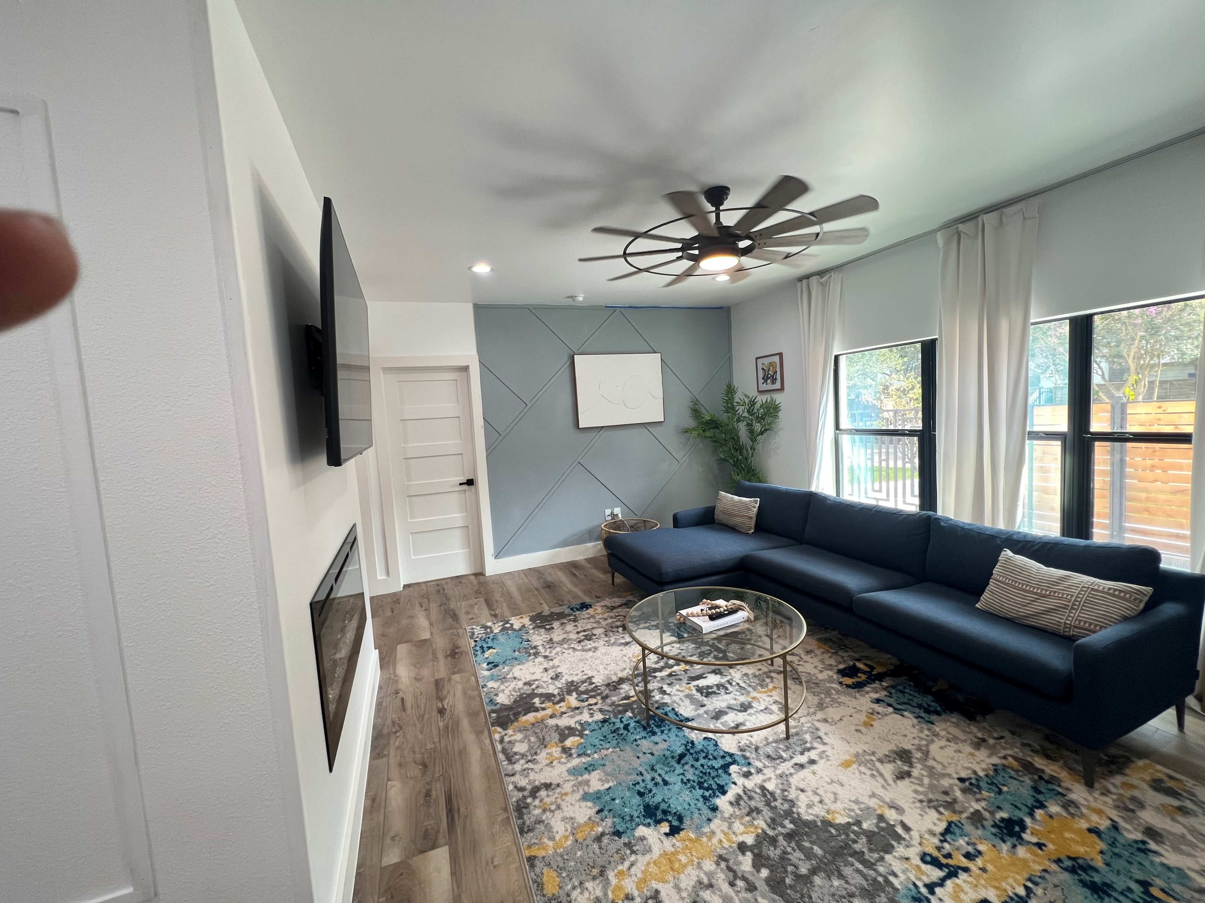 The image shows a modern living room featuring a blue sectional sofa, a round coffee table on an area rug, a flat-screen TV mounted on the wall, and large windows allowing natural light.