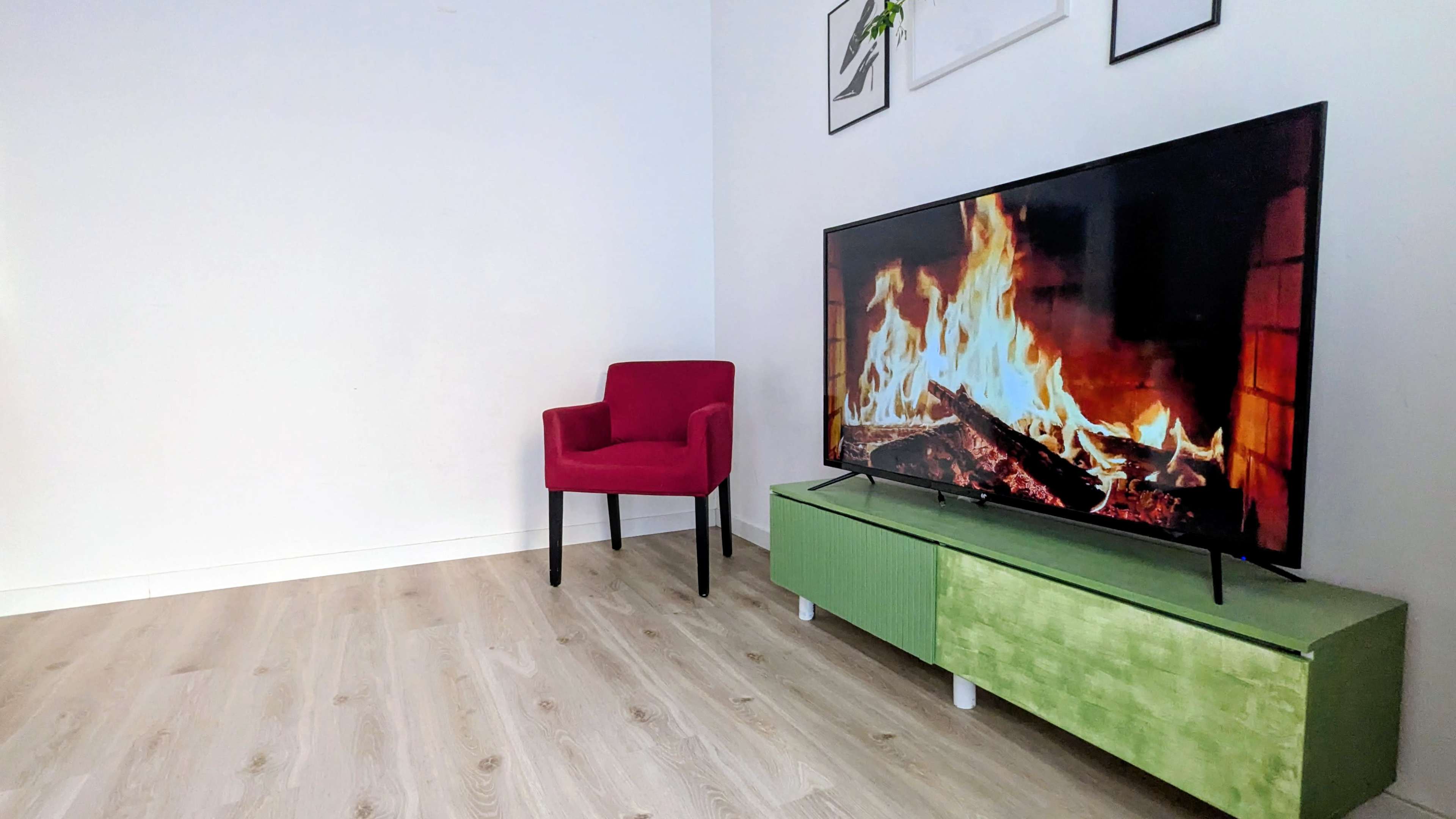 A red chair is positioned next to a green TV stand displaying a fire-themed image on a large flat-screen television, set in a minimalist room with light wooden flooring.