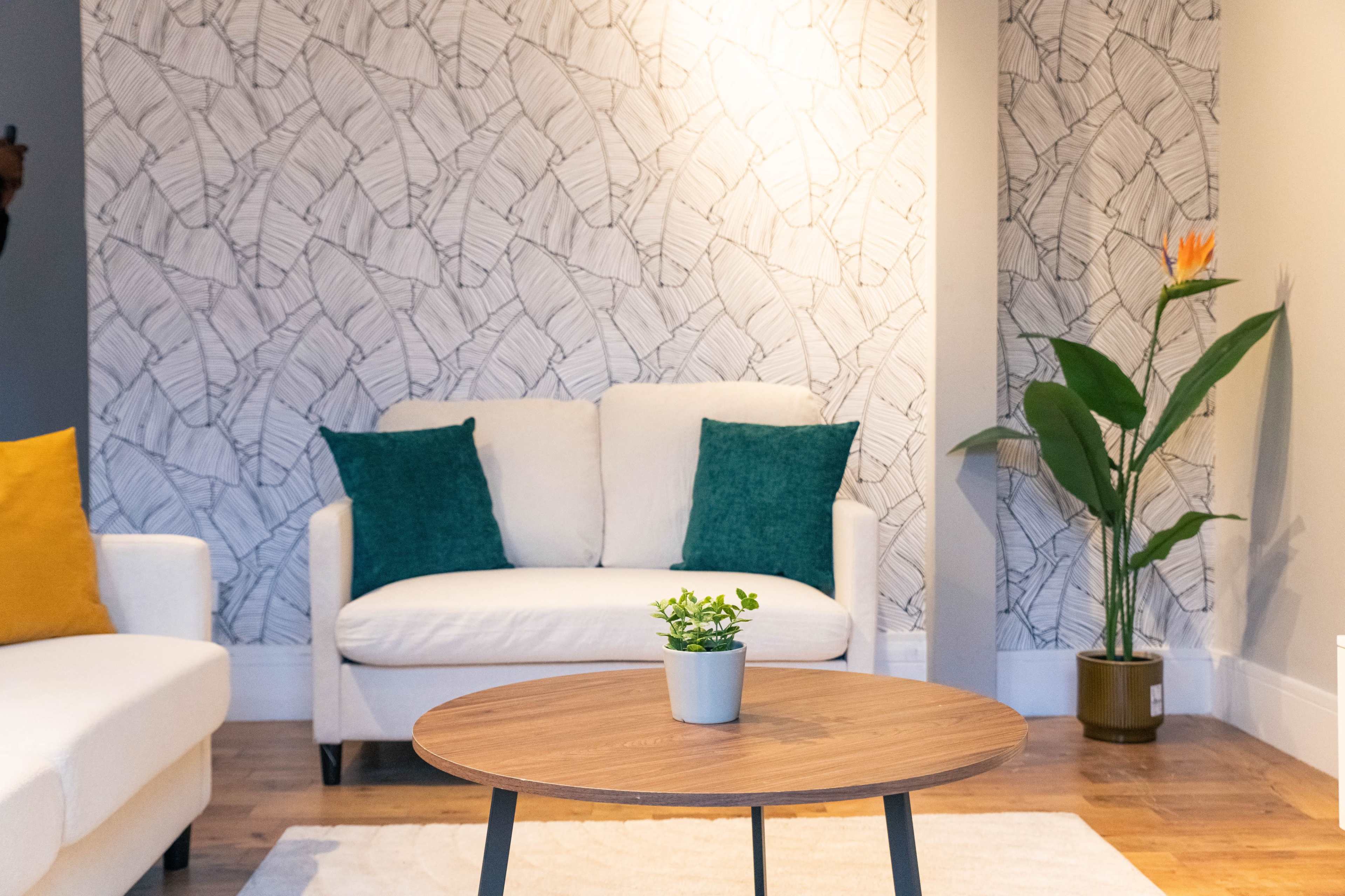 The image shows a modern living room with a white couch, a round wooden coffee table, and a decorative plant in a corner against a patterned wall.