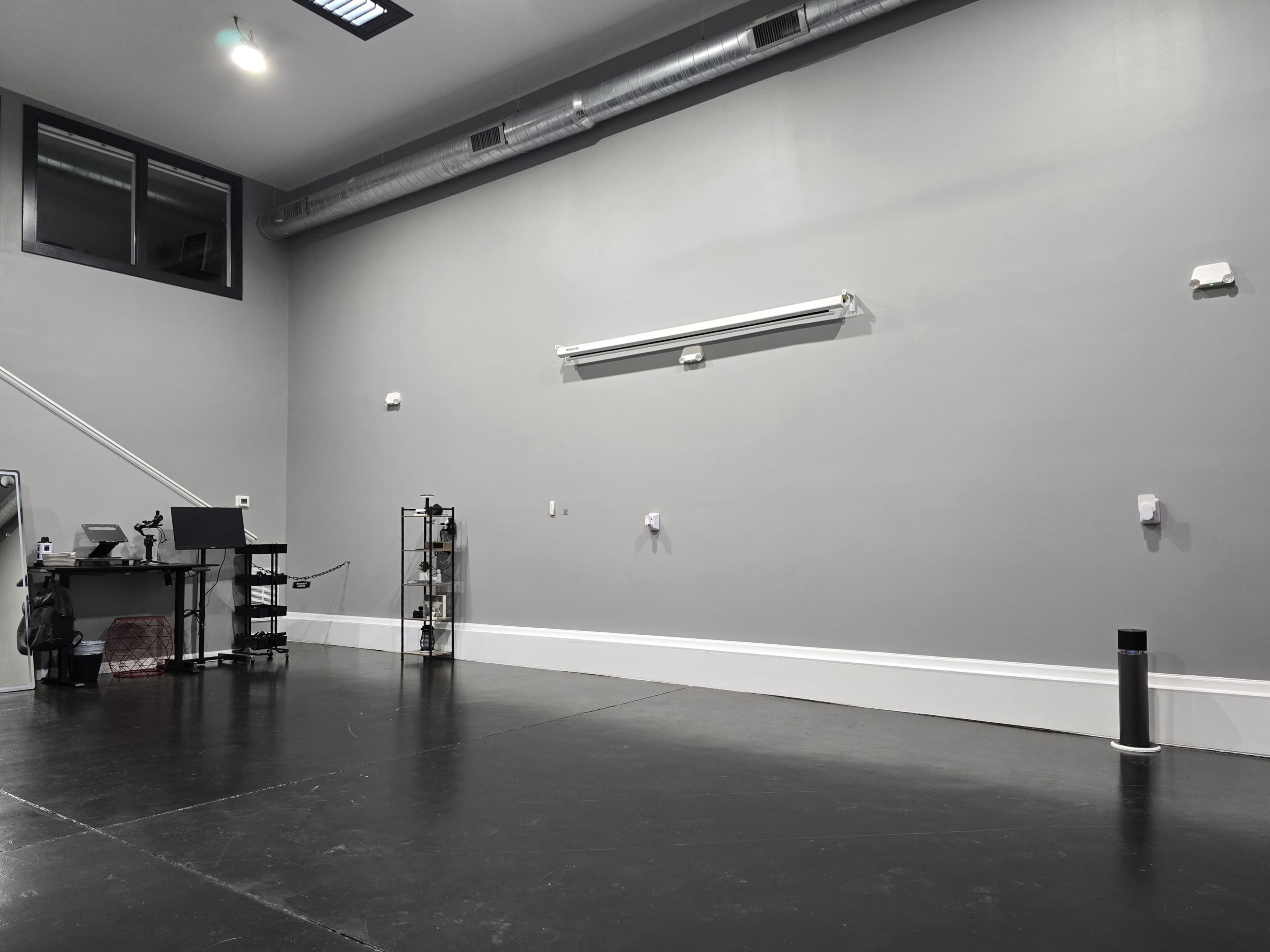 The image shows a blank, spacious interior with a gray wall, a black floor, and minimal furnishings, including a table with equipment and a shelving unit.