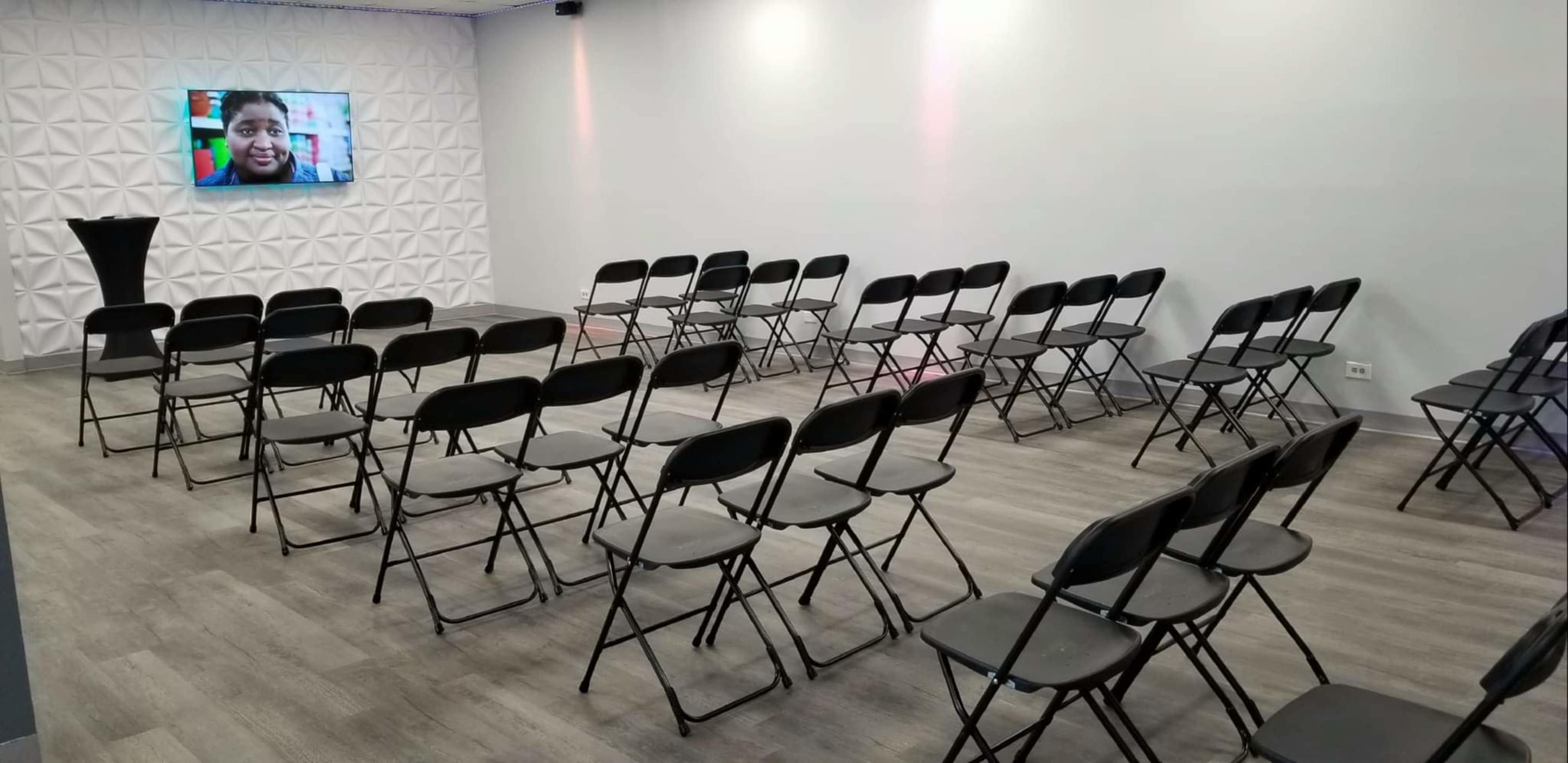 A room with neatly arranged black folding chairs facing a wall featuring a screen displaying a person's image.