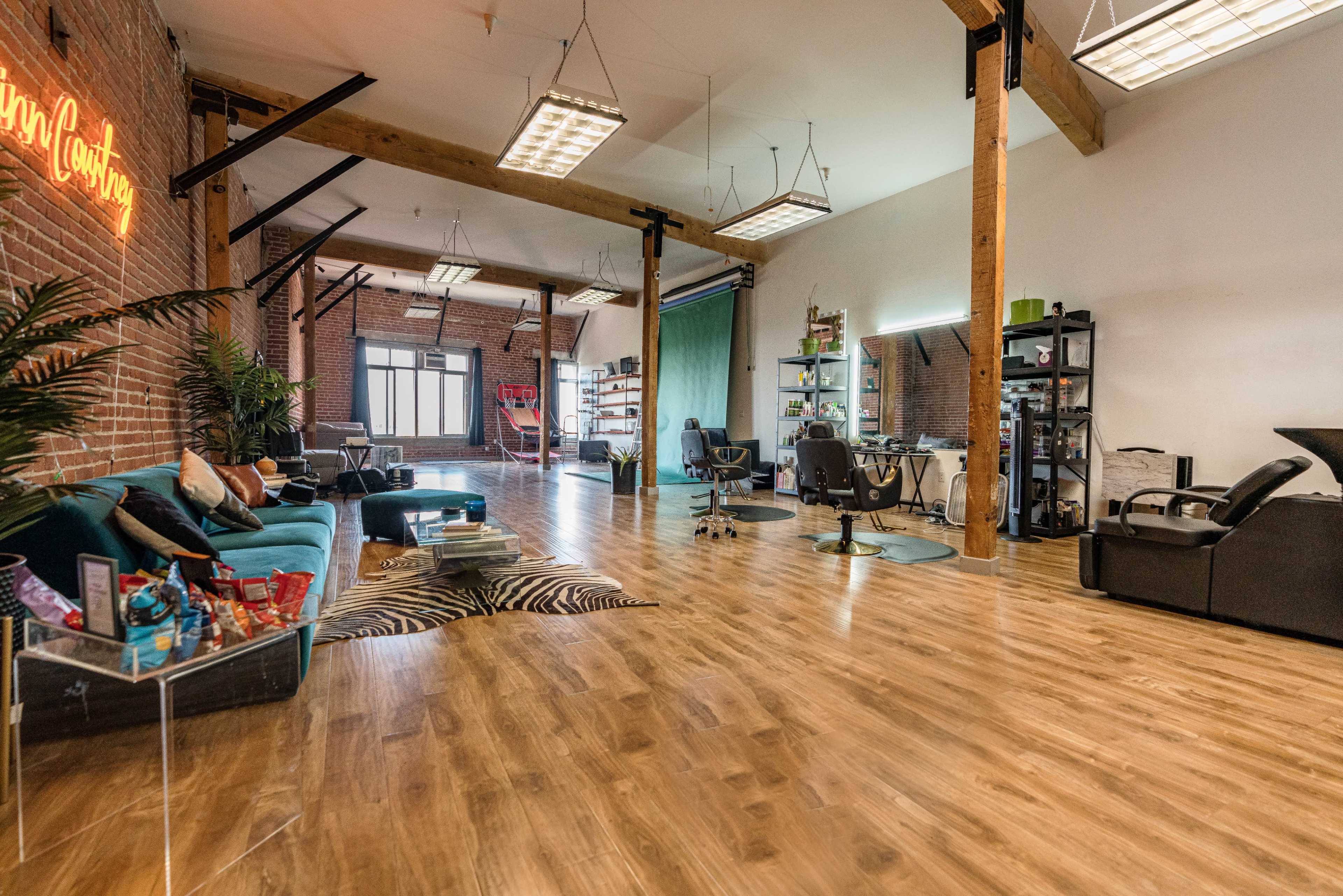 A spacious, modern studio with wooden flooring, large windows, and a mix of seating areas, including a teal sofa and several salon-style chairs.