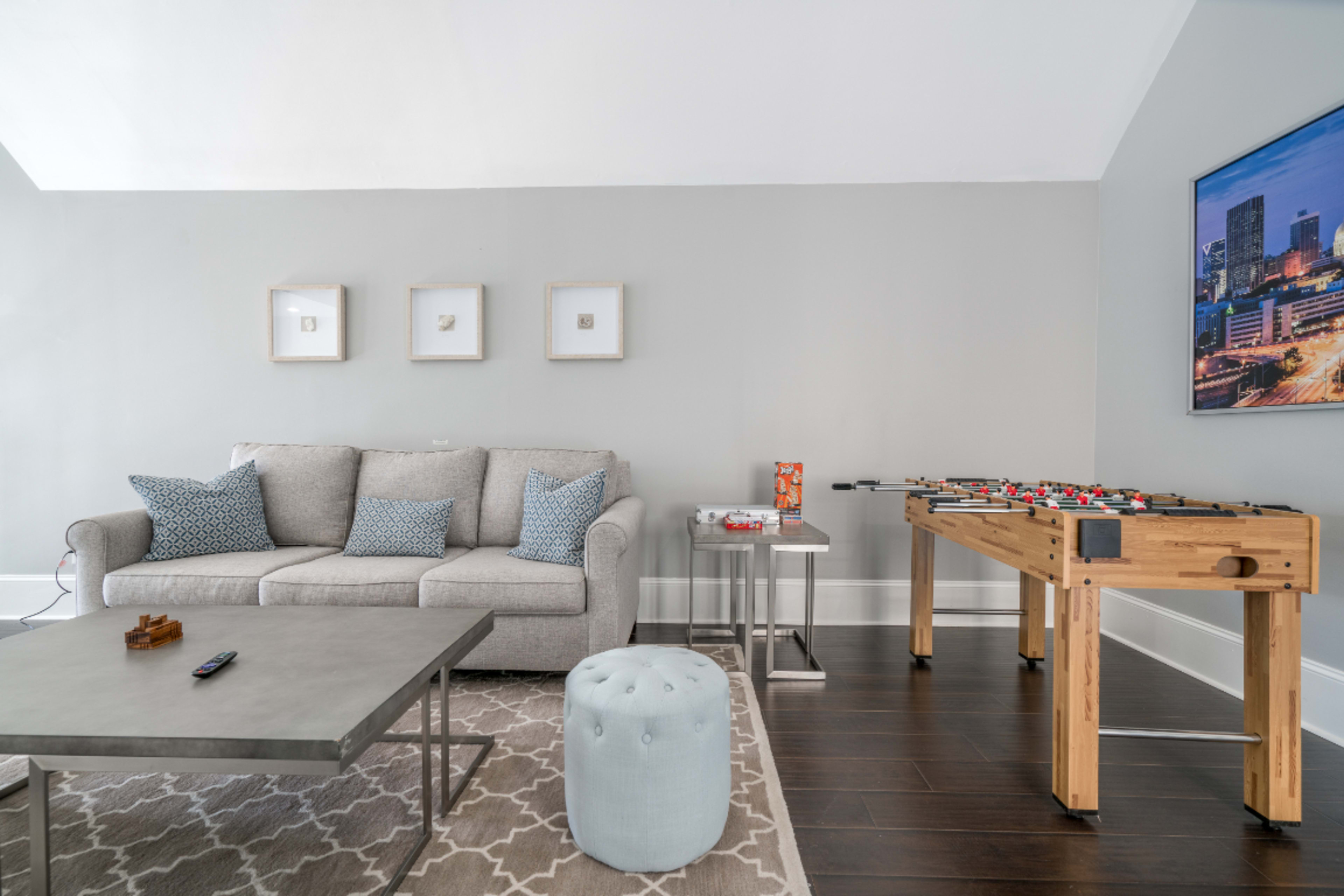The image shows a living room with a gray sofa, a coffee table, and a foosball table, situated against a light gray wall.