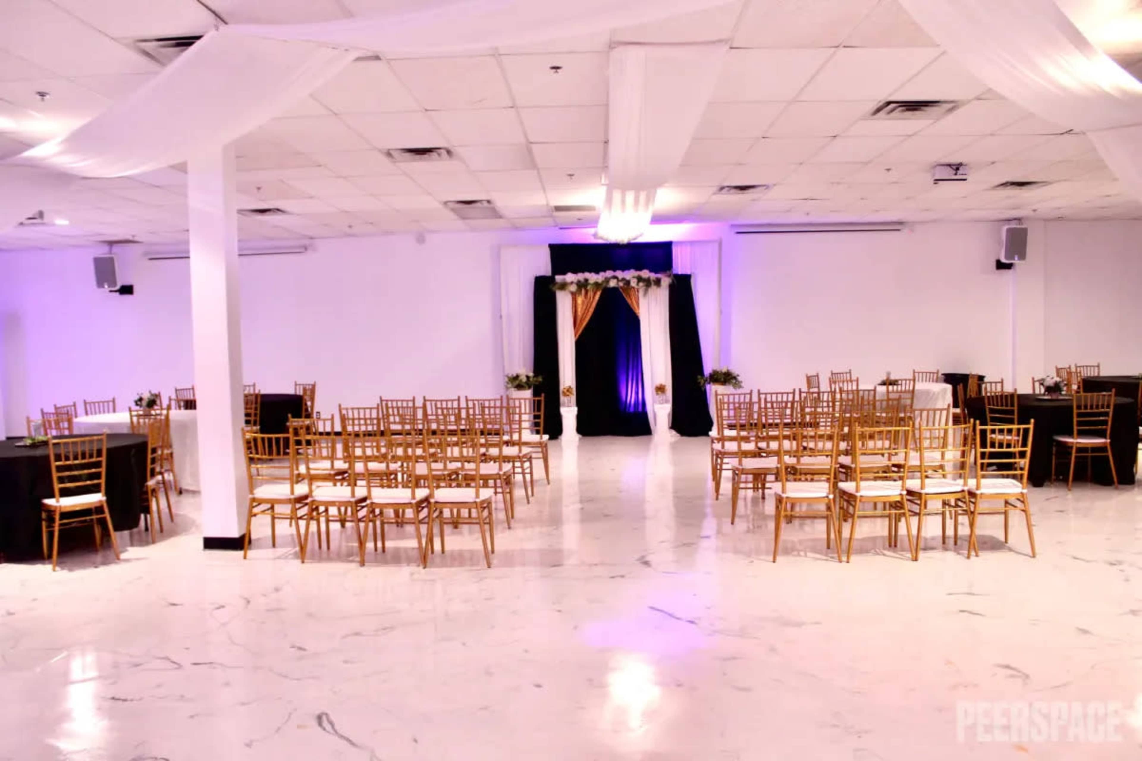 An event space arranged with gold chairs and tables set for a gathering, featuring a decorative backdrop at one end.