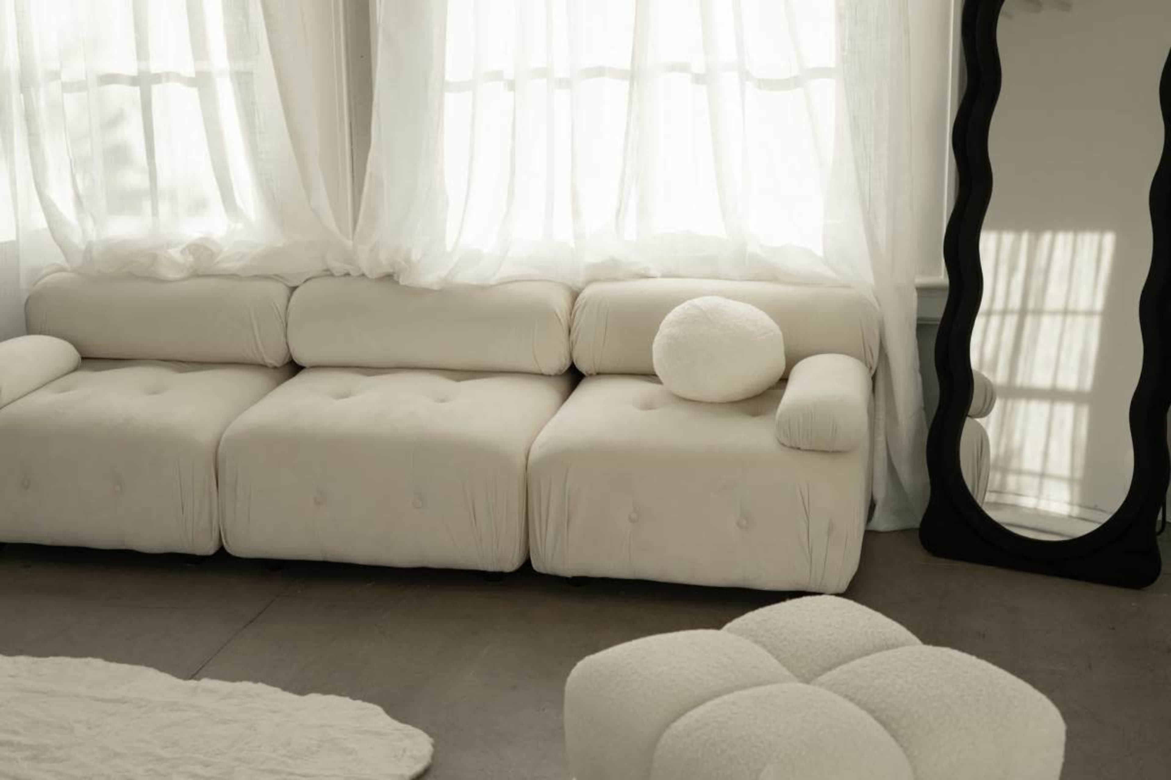 A light-colored, modern sofa with tufted upholstery is positioned near large windows, accompanied by an oval mirror and a small, round ottoman.