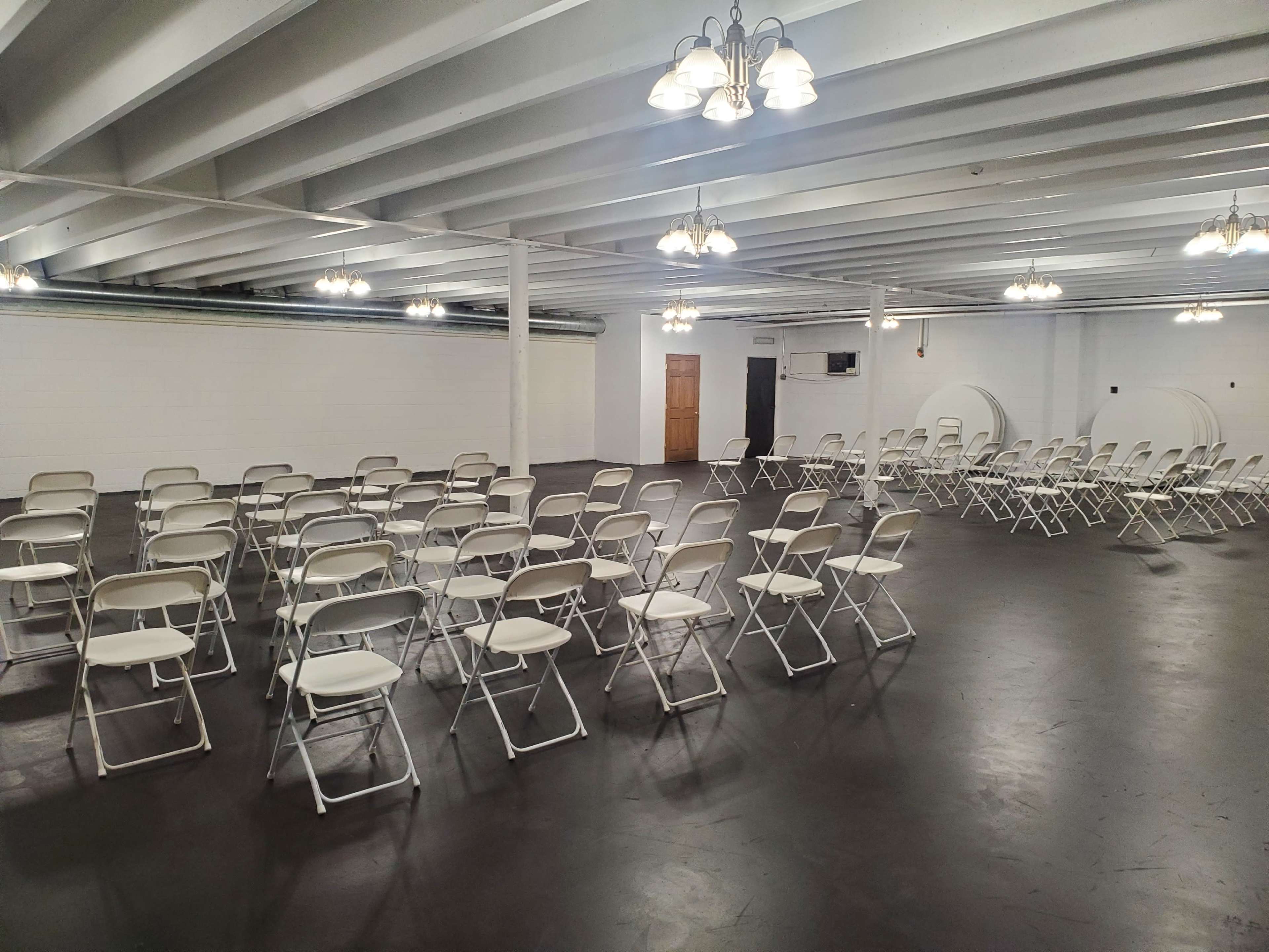 A large, empty room arranged with multiple rows of white folding chairs facing a central area, illuminated by overhead lights.
