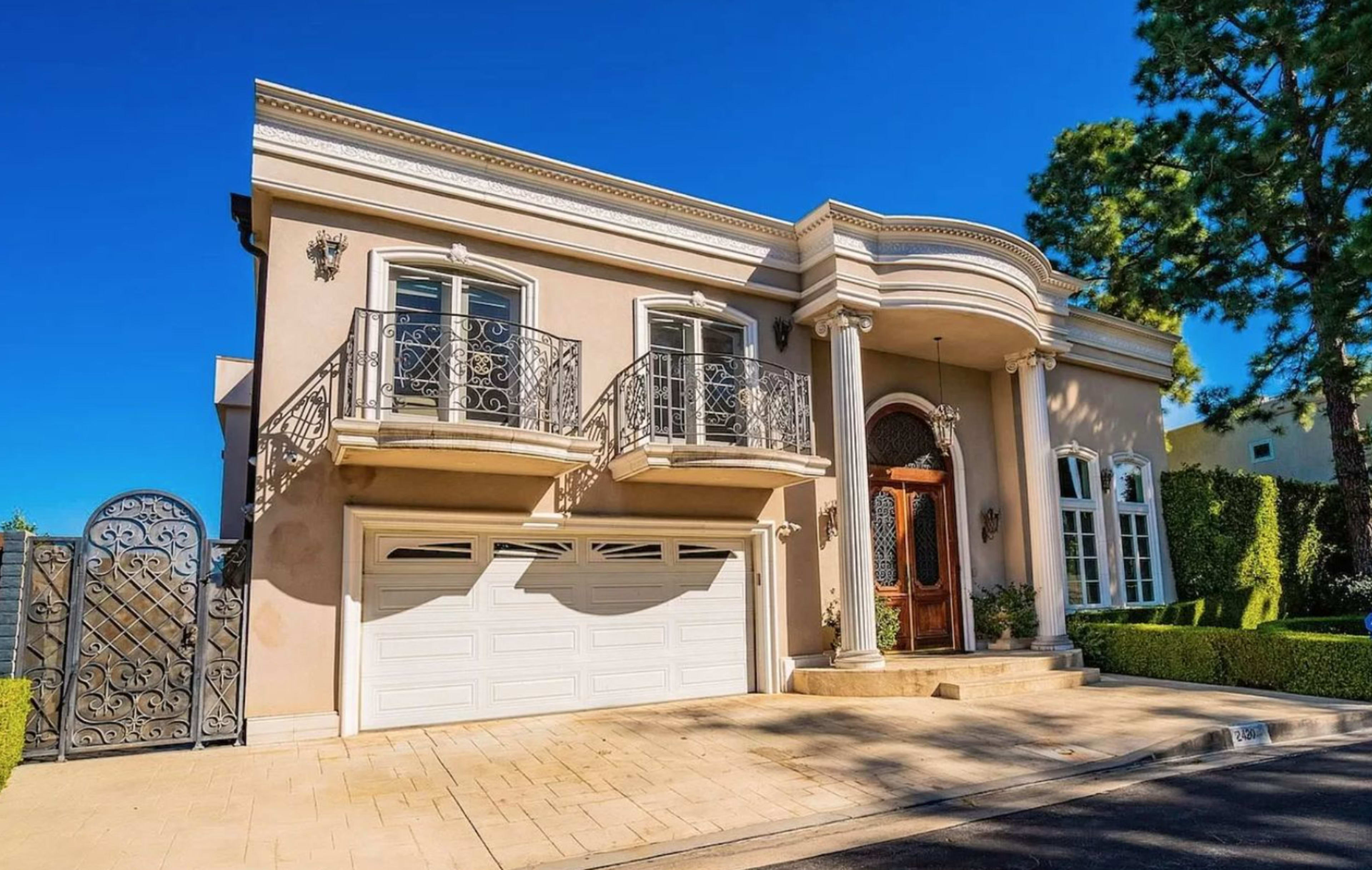 The image shows a large, modern stucco house with a decorative entrance, two balconies, and a three-car garage, set in a well-manicured environment.