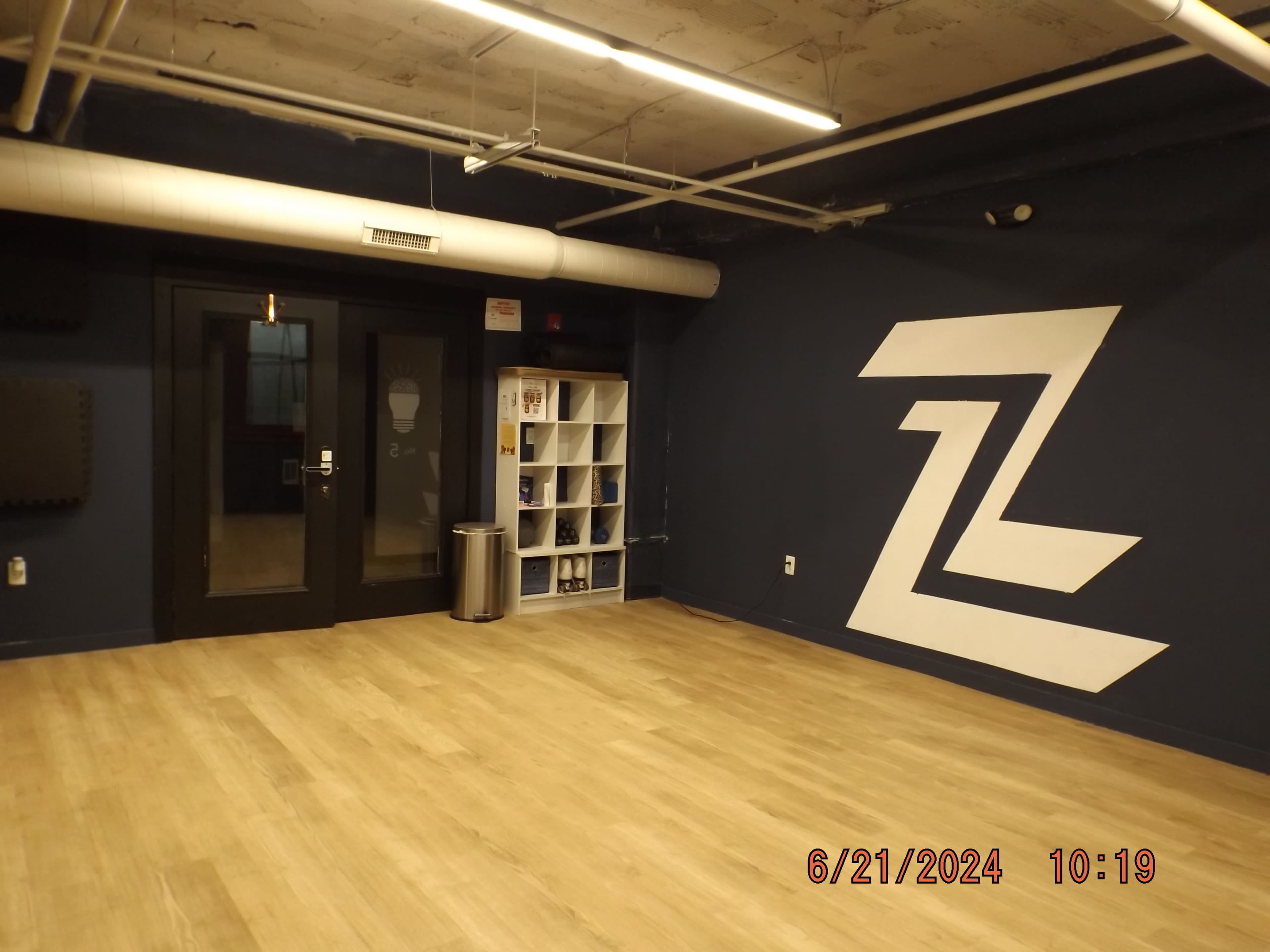 The image shows a spacious room with a wooden floor, featuring a dark blue wall with a large white logo and a glass door leading to another area.