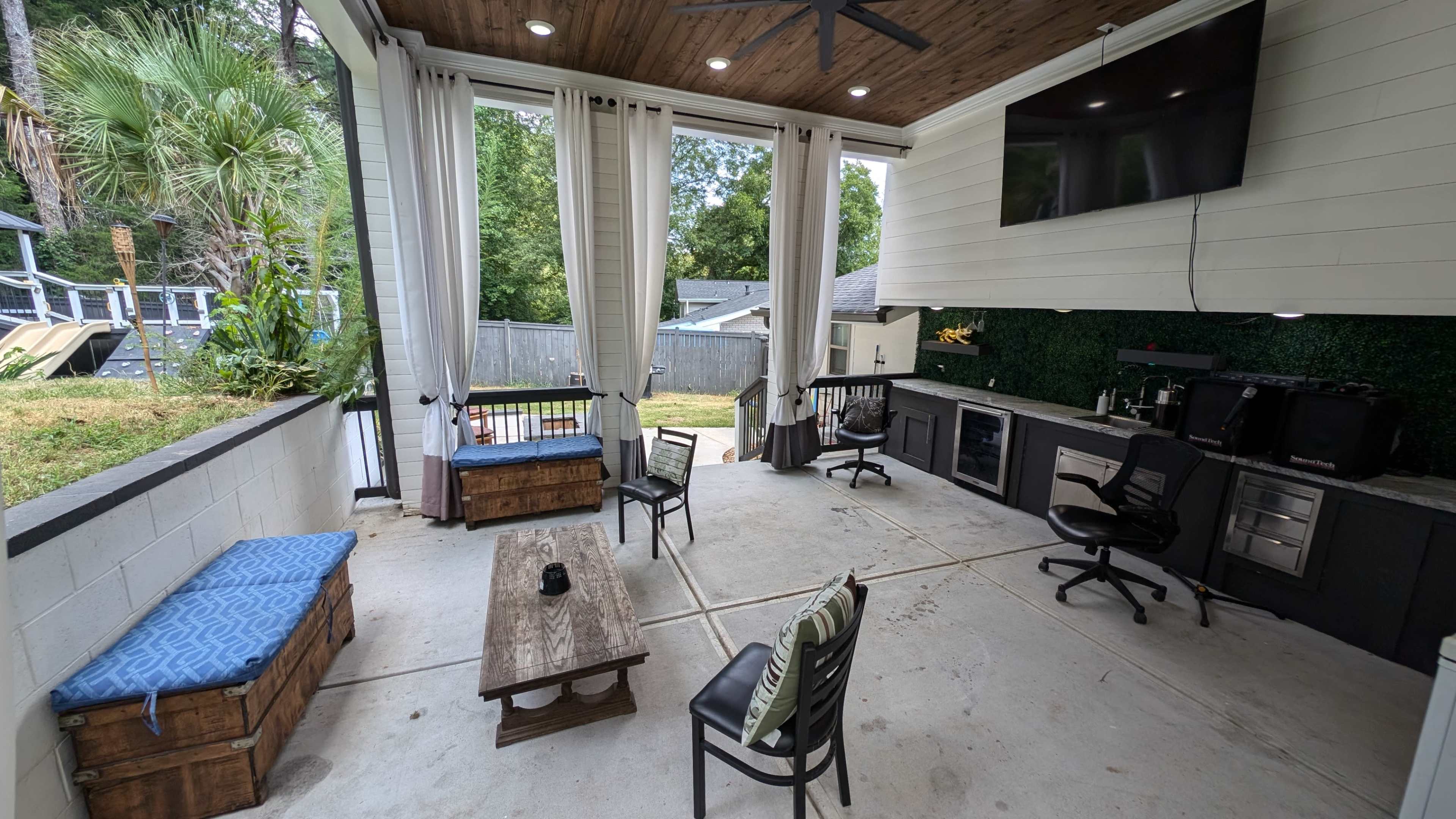 An outdoor living space featuring a patio with seating, a wooden table, a bar area with a countertop, and large curtains along the sides.