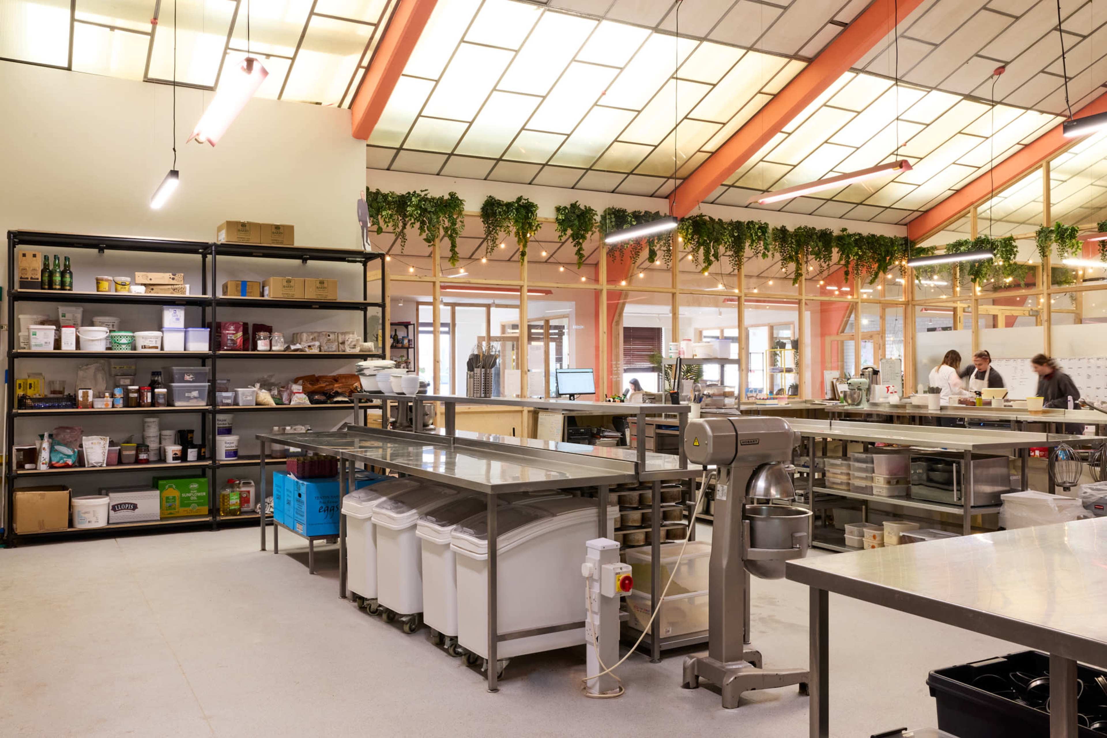 The image shows a spacious kitchen area with stainless steel worktables, shelves filled with various culinary supplies, and large windows letting in natural light.