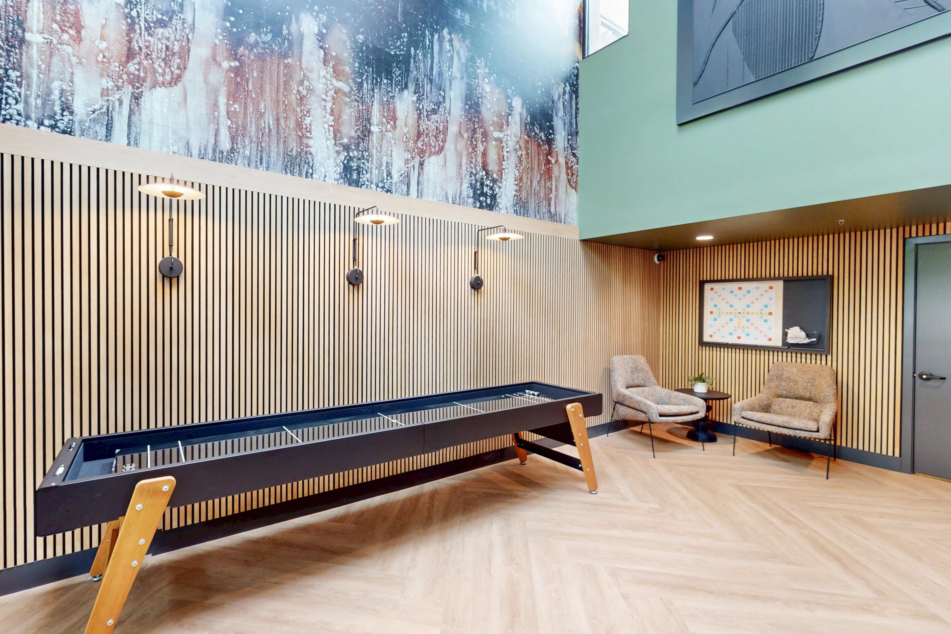The image shows a modern indoor space with wooden paneling, a shuffleboard table, and two armchairs beside a wall displaying artwork.