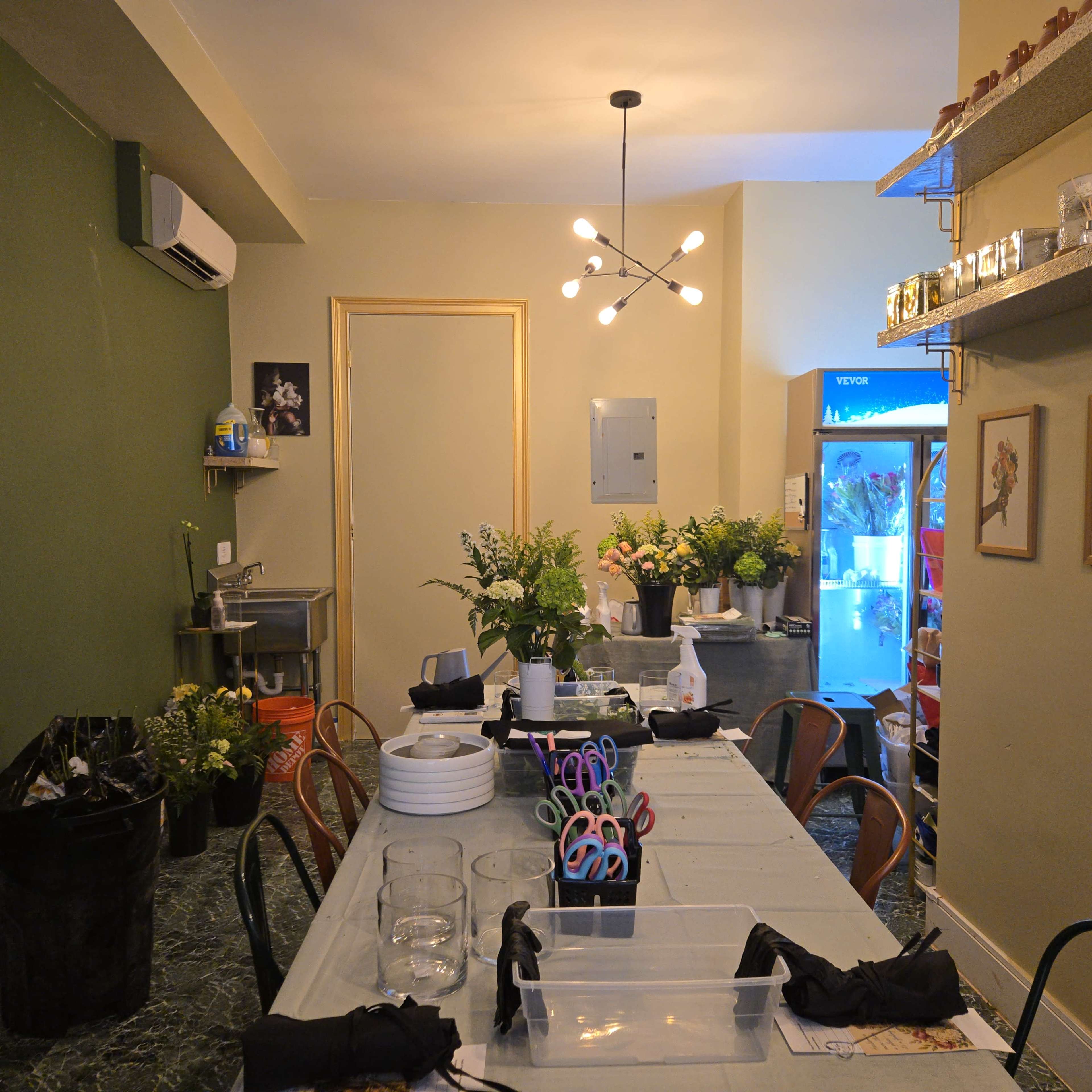 The image shows a cozy workshop space with a long table set for an activity, surrounded by plants, supplies like scissors, and a visible refrigerator in the background.