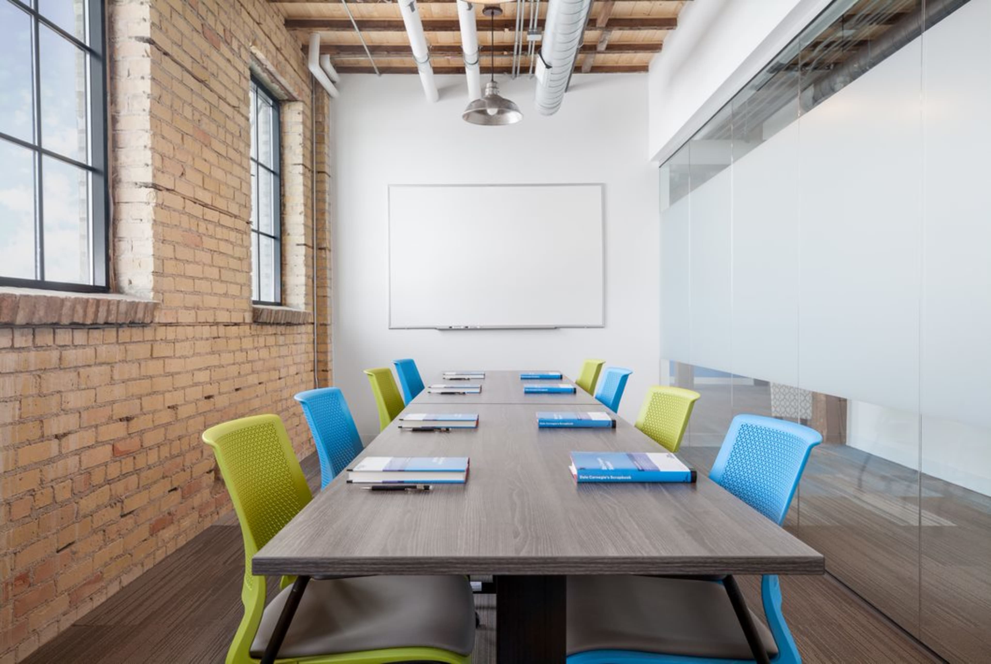 A modern conference room features a long table with neatly arranged folders and chairs in blue and green along the sides, and a whiteboard is mounted on the wall.